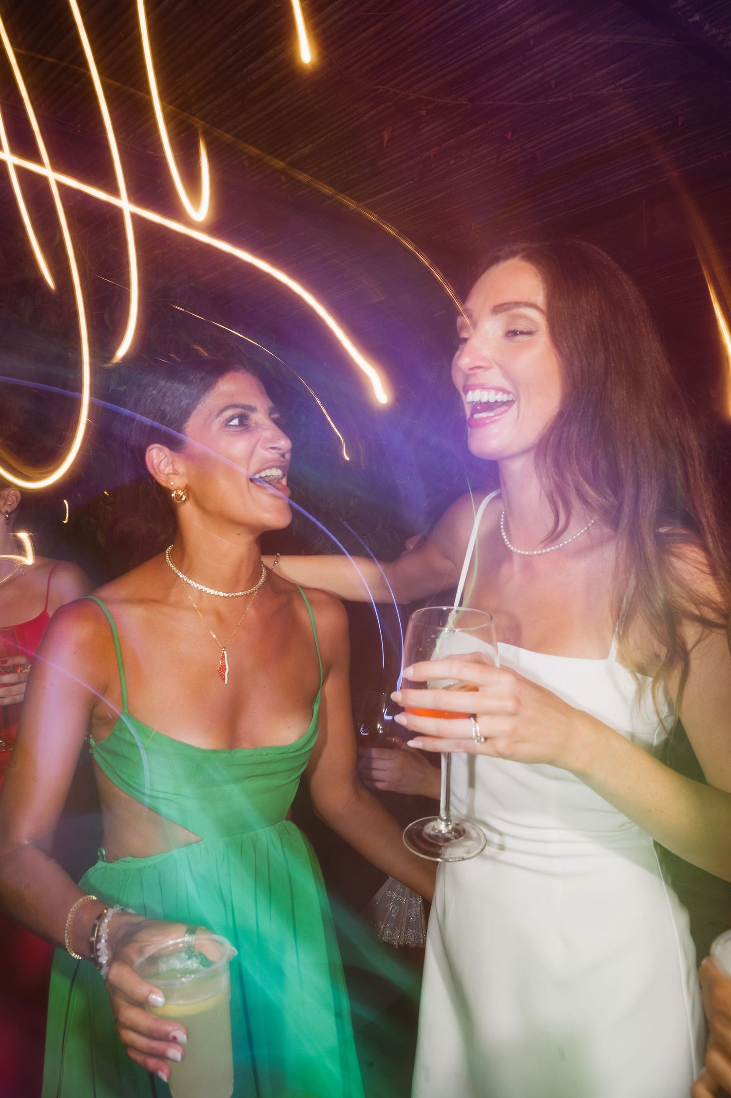 Two people in green and white dresses dance and laugh together at a party with streaks of light in the background.