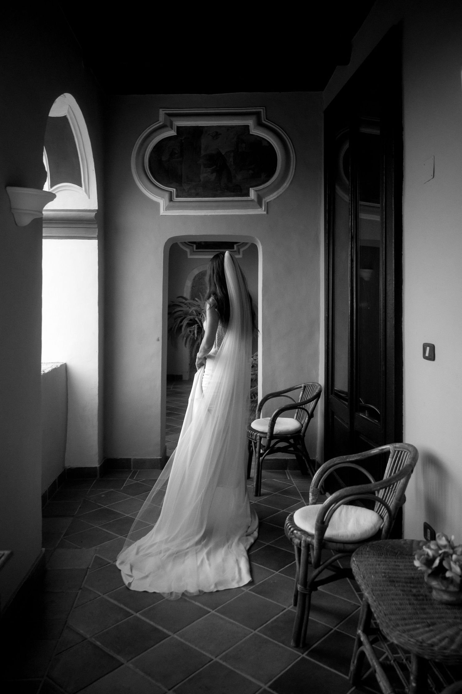 A flowing white wedding dress hangs elegantly in a doorway of an ornate hallway with rattan chairs and dramatic lighting.