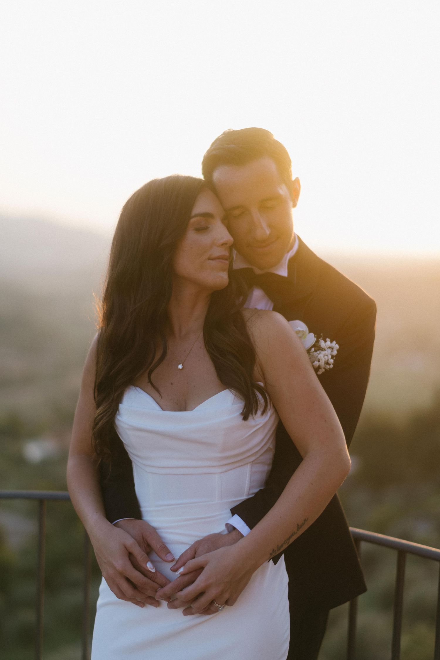 A couple shares an intimate sunset moment on a balcony in formal wedding attire.