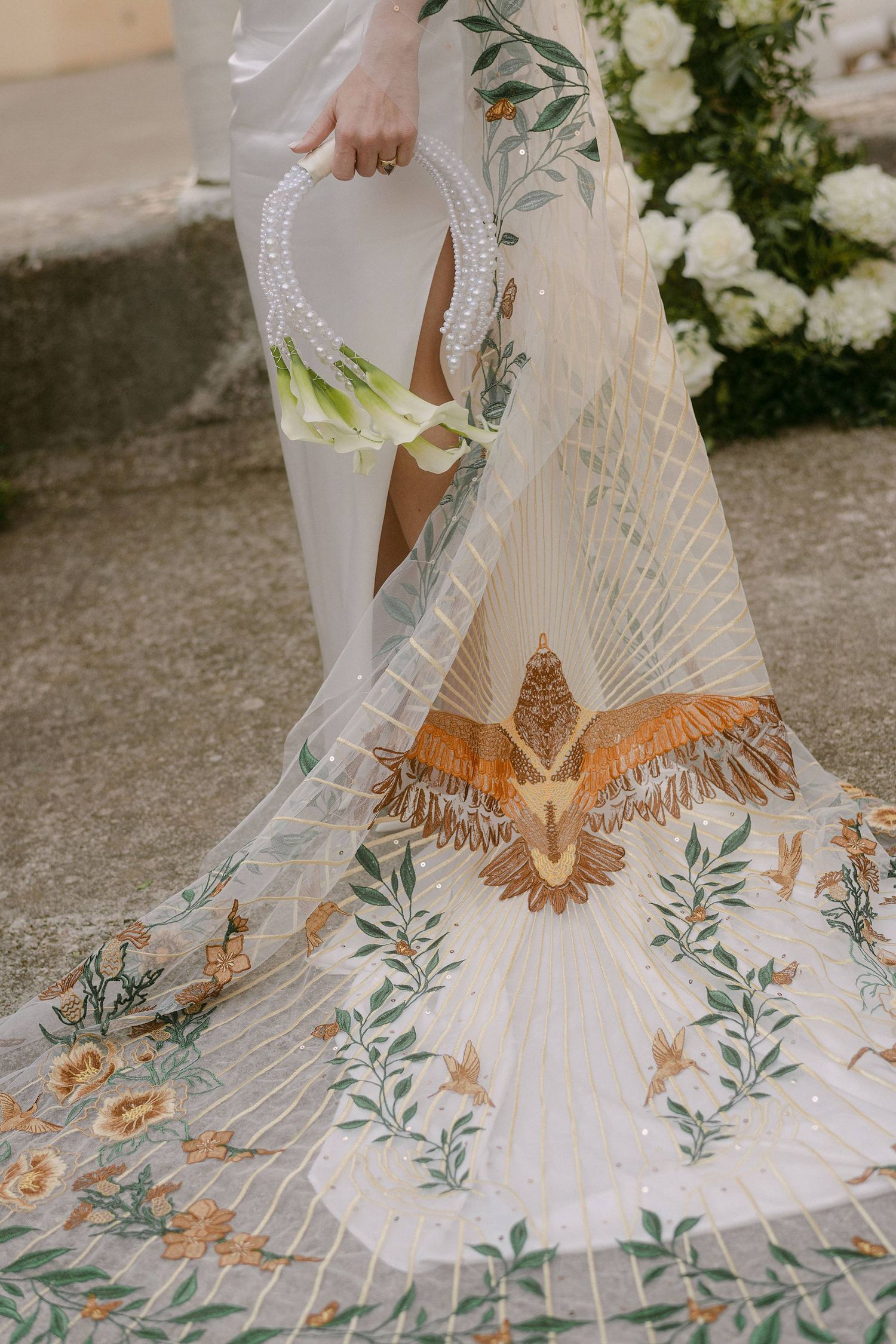 Intricate wedding dress with golden eagle embroidery and floral designs trails along stone floor among white flowers.