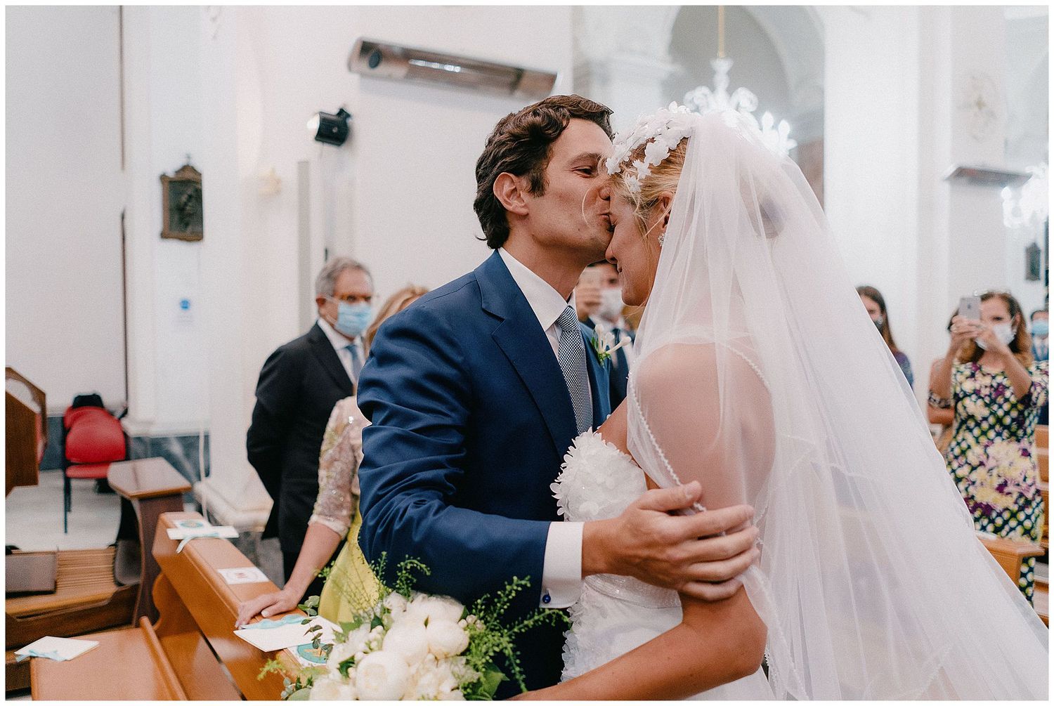 Two people share a romantic kiss at the altar during their wedding ceremony inside a church while guests look on.