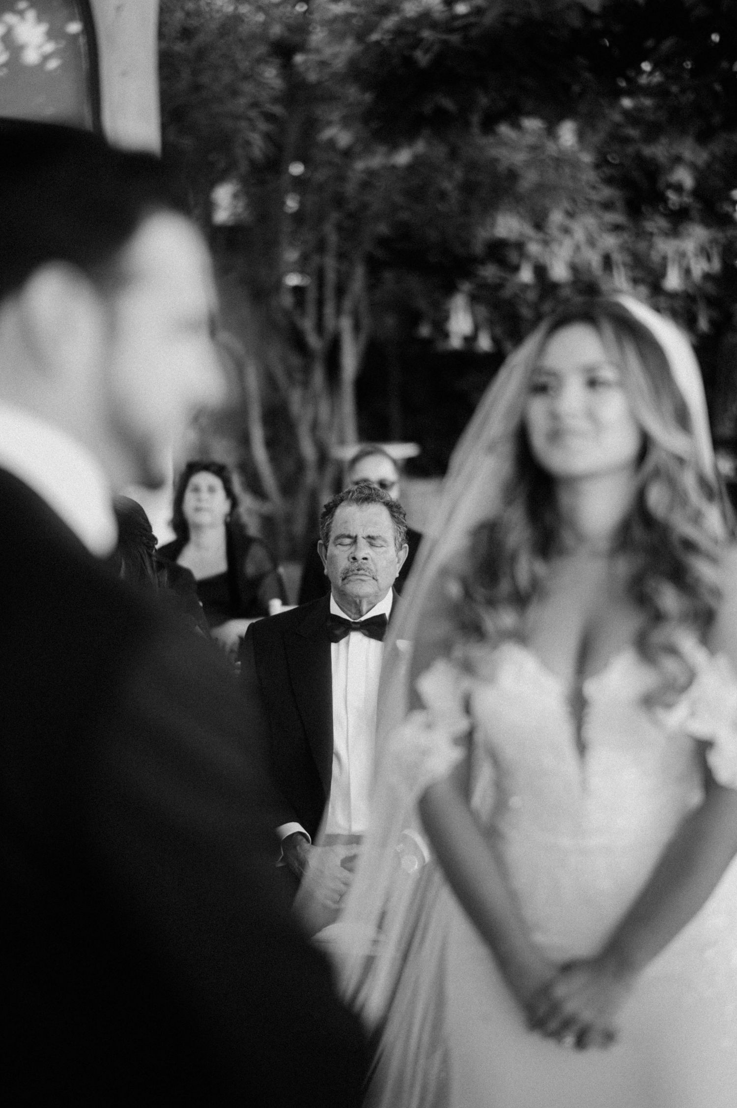 Black and white wedding ceremony photo captures a candid moment with guests and a couple in formal attire.