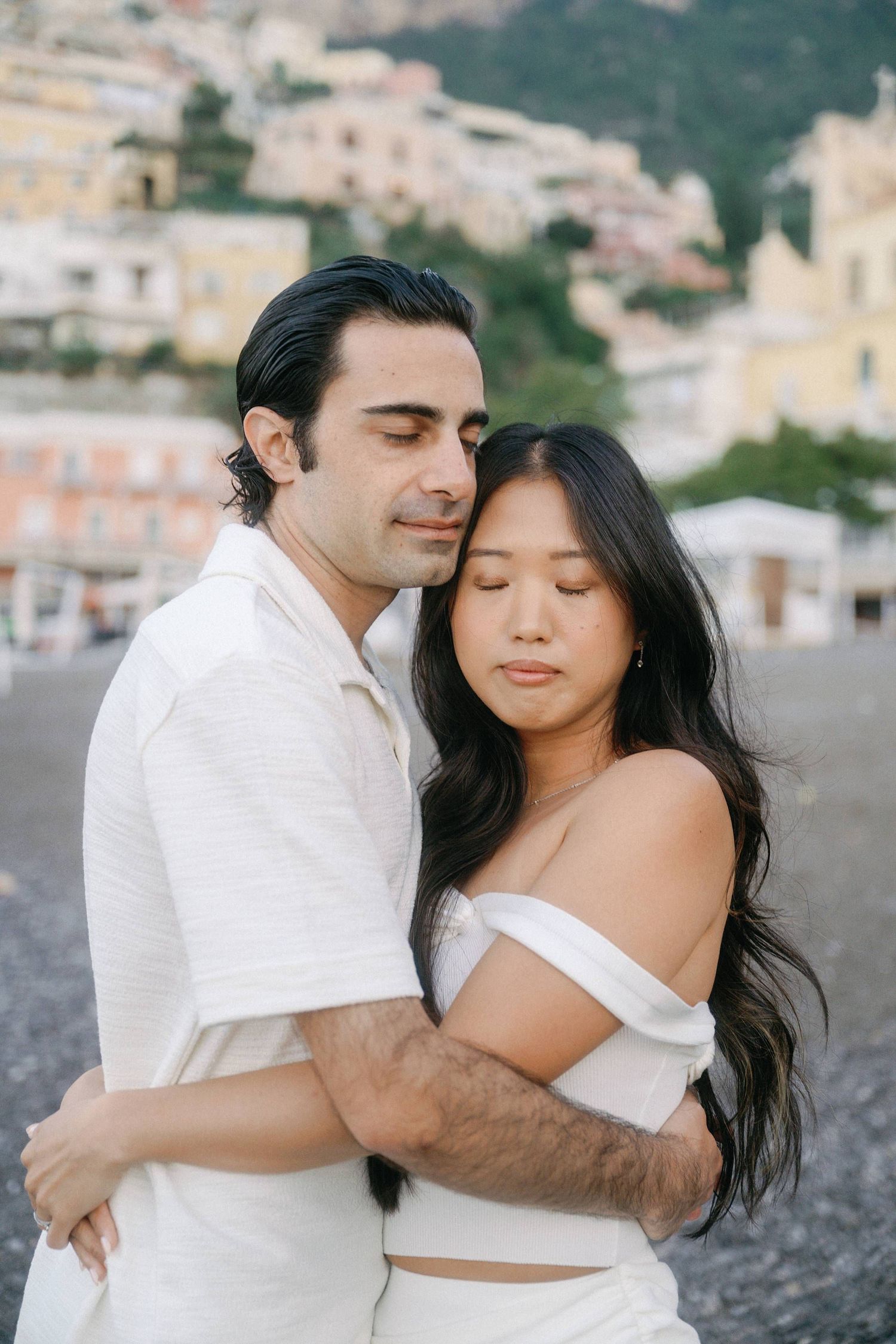 A romantic couple embraces on a pebble beach with the colorful buildings of Positano, Italy in the background.