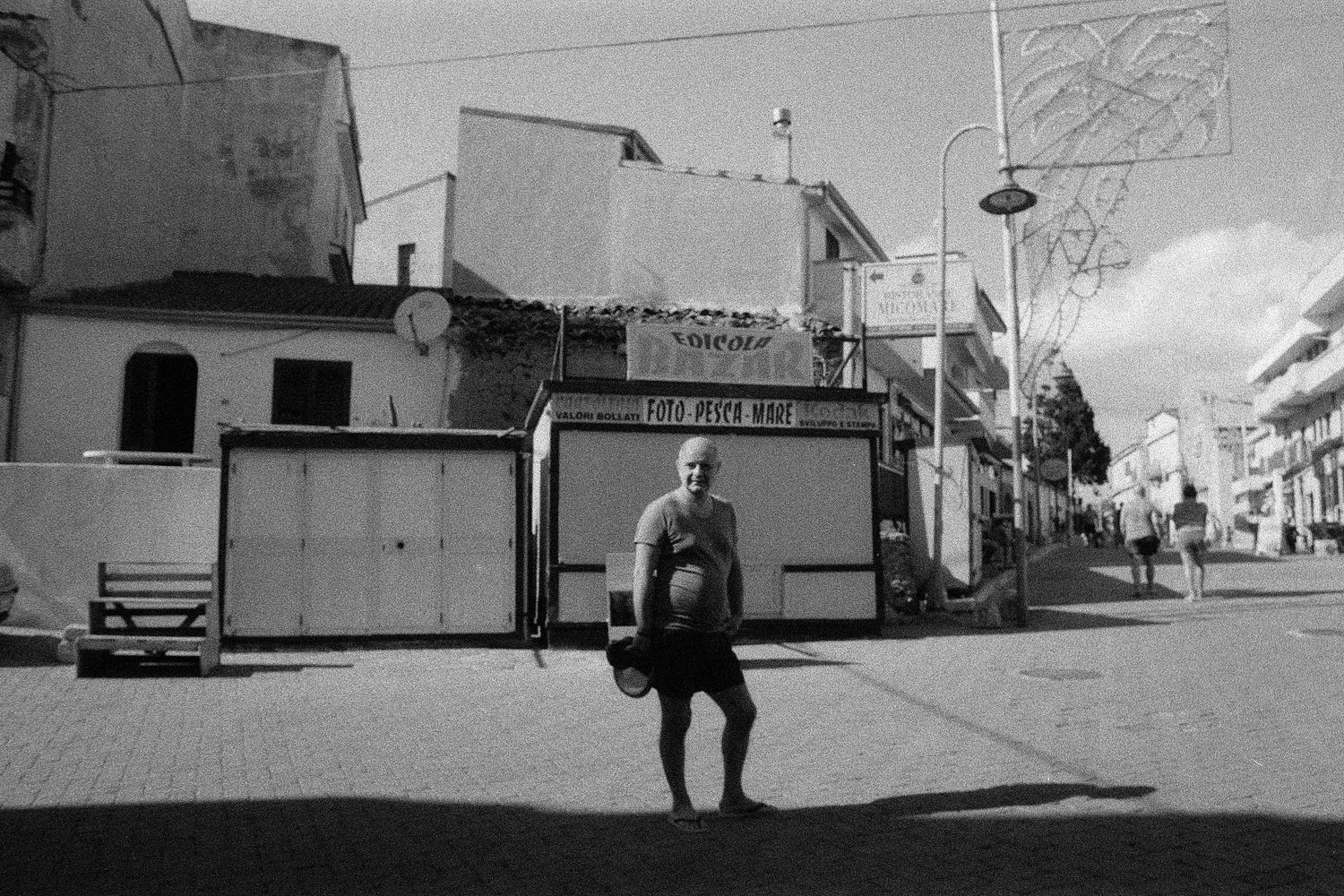A black and white street photograph shows a person sitting on a stool in front of closed businesses and storefronts.