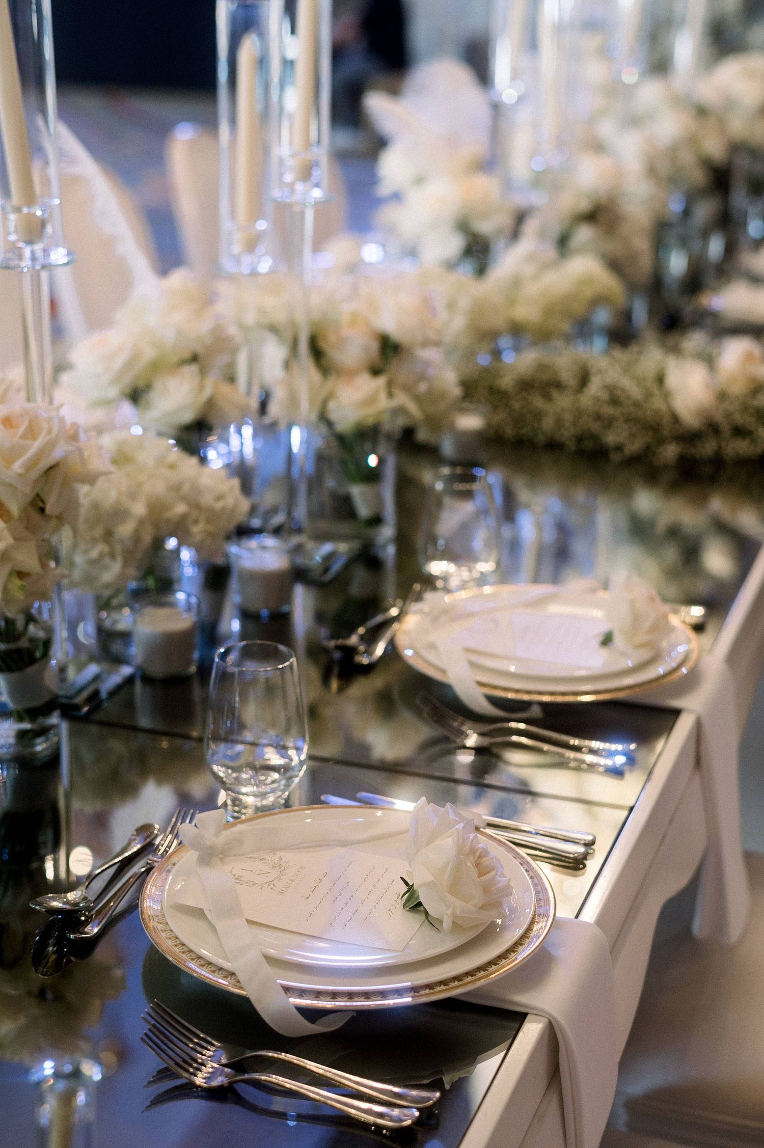 Elegant table setting with white plates, silverware, and delicate floral centerpieces on a reflective surface.