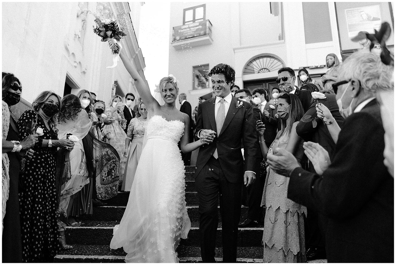 A joyful couple walks down church steps while wedding guests shower them with rice in a black and white photograph.