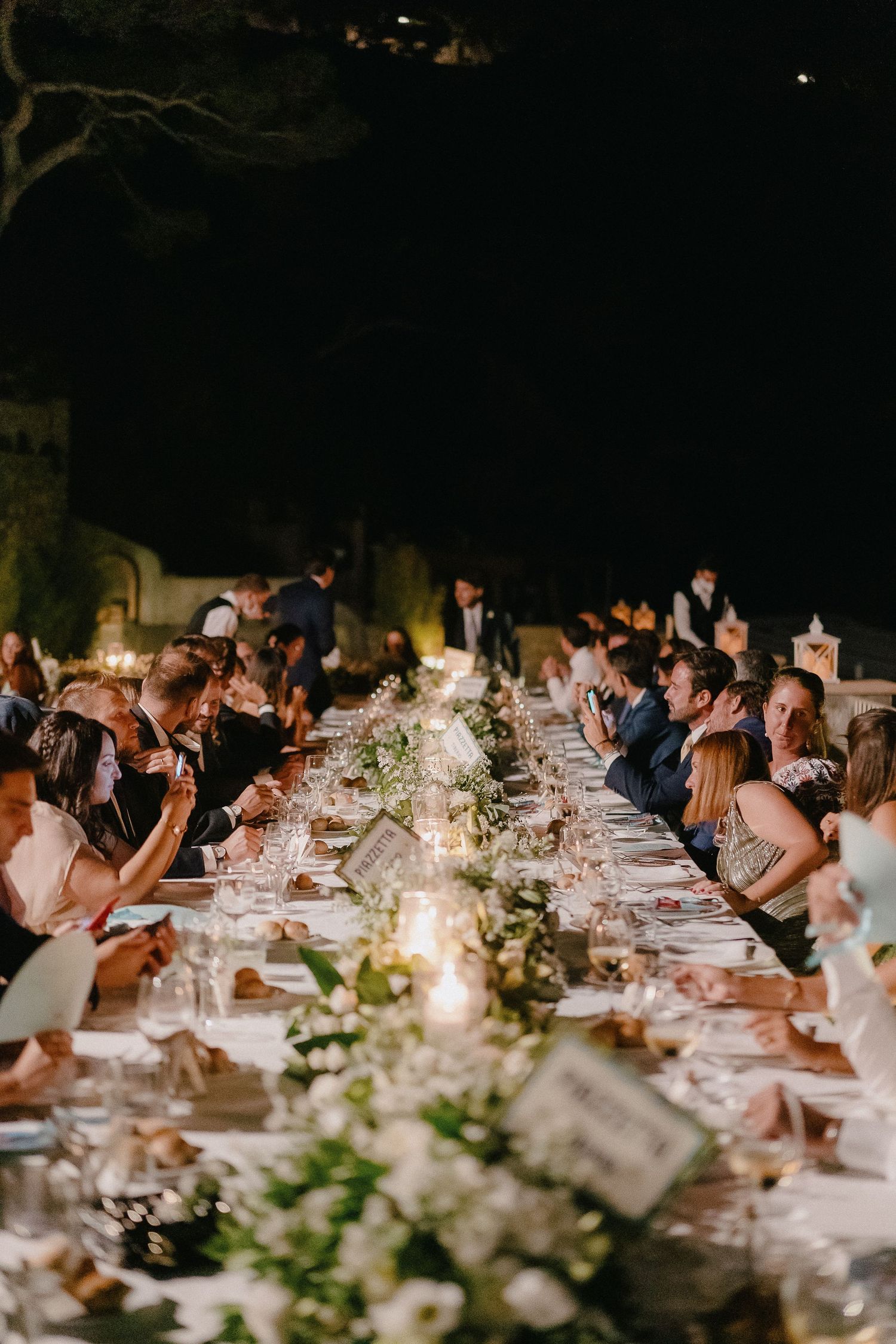 Guests seated at an elegant long dining table with candlelight and floral decorations at an outdoor evening reception.