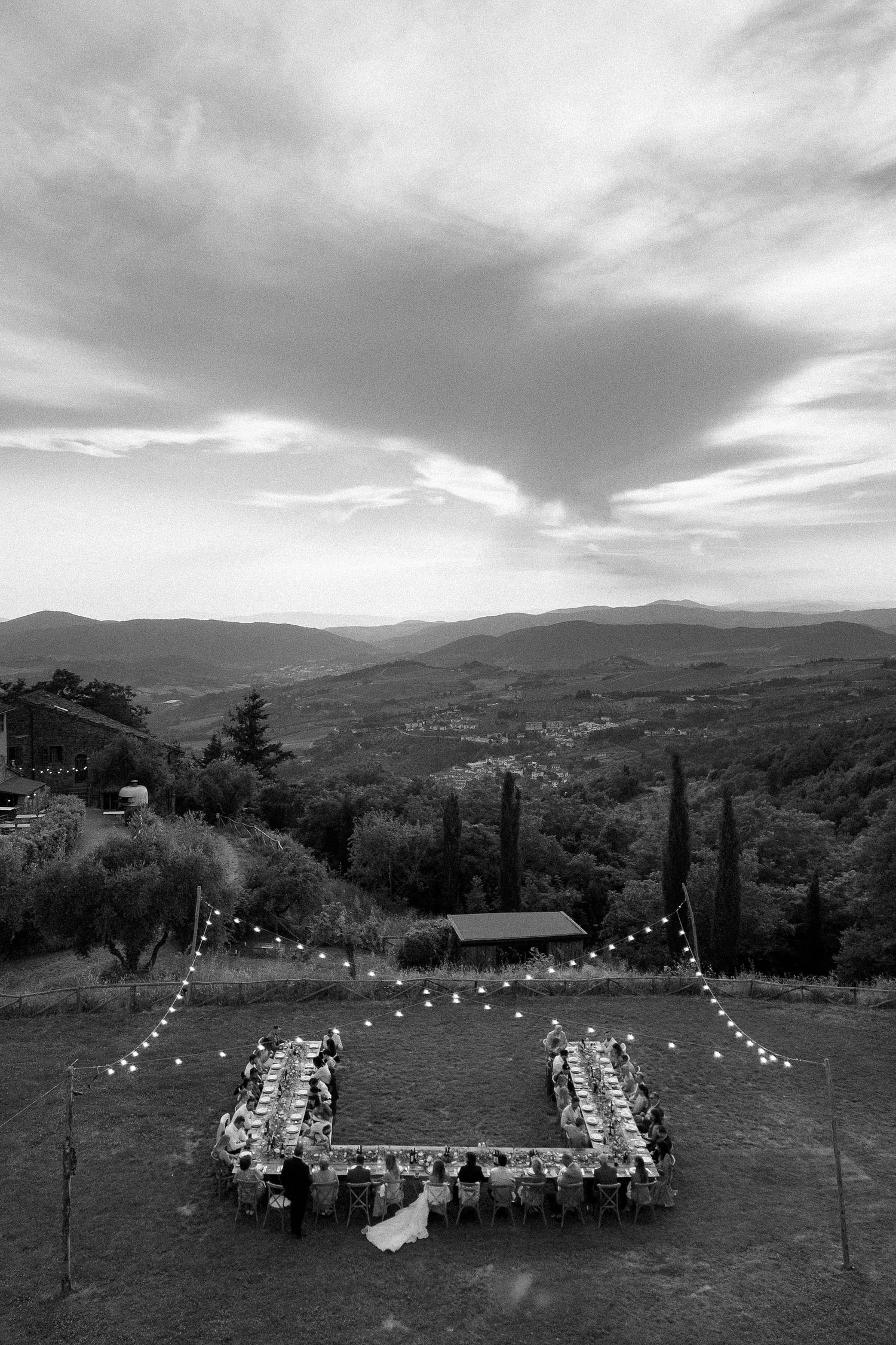 Aerial black and white photo of an outdoor wedding reception with string lights set against rolling Tuscan hills.