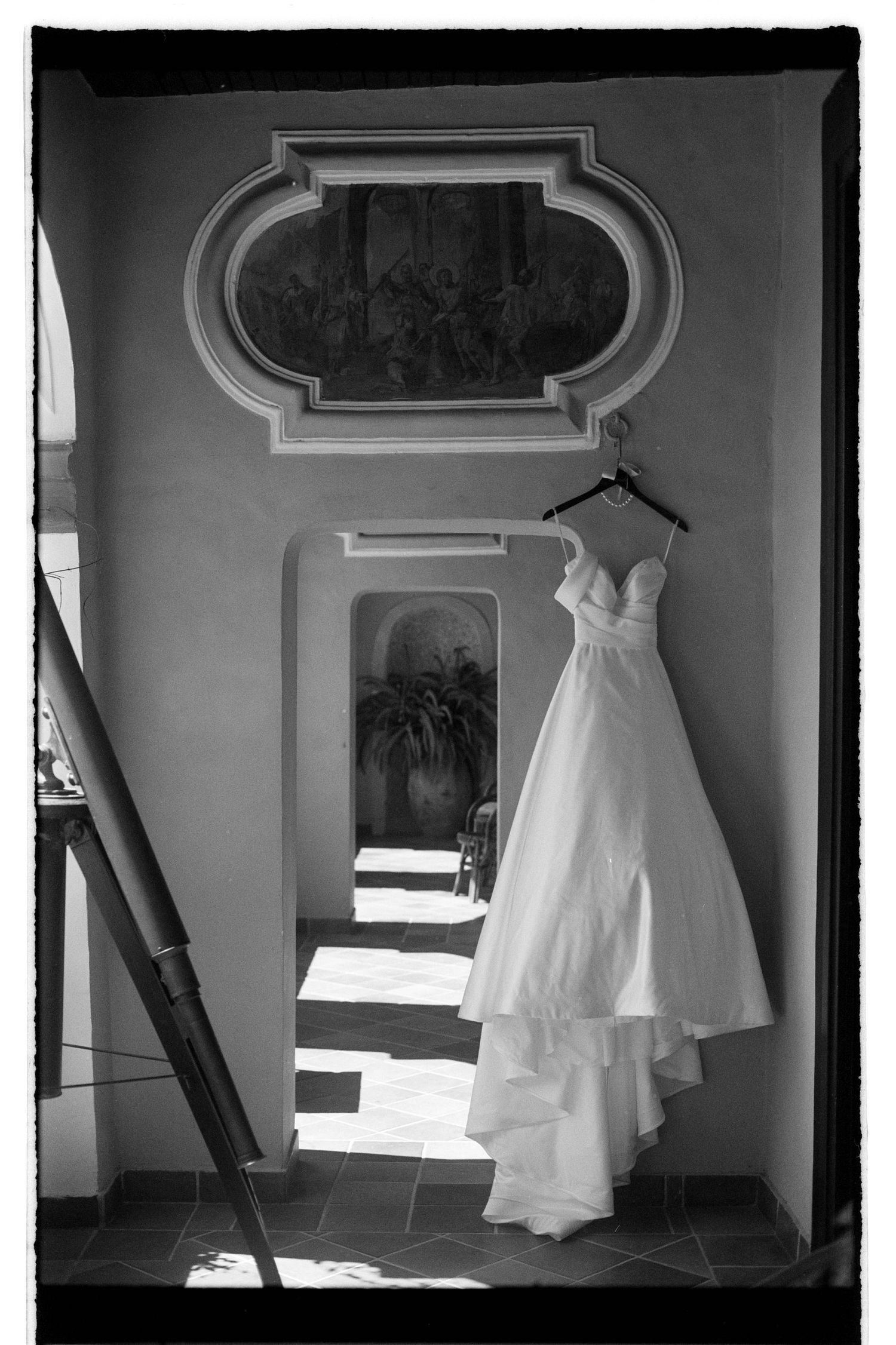 A white wedding dress hangs in a dimly lit room with architectural details and window light casting dramatic shadows.