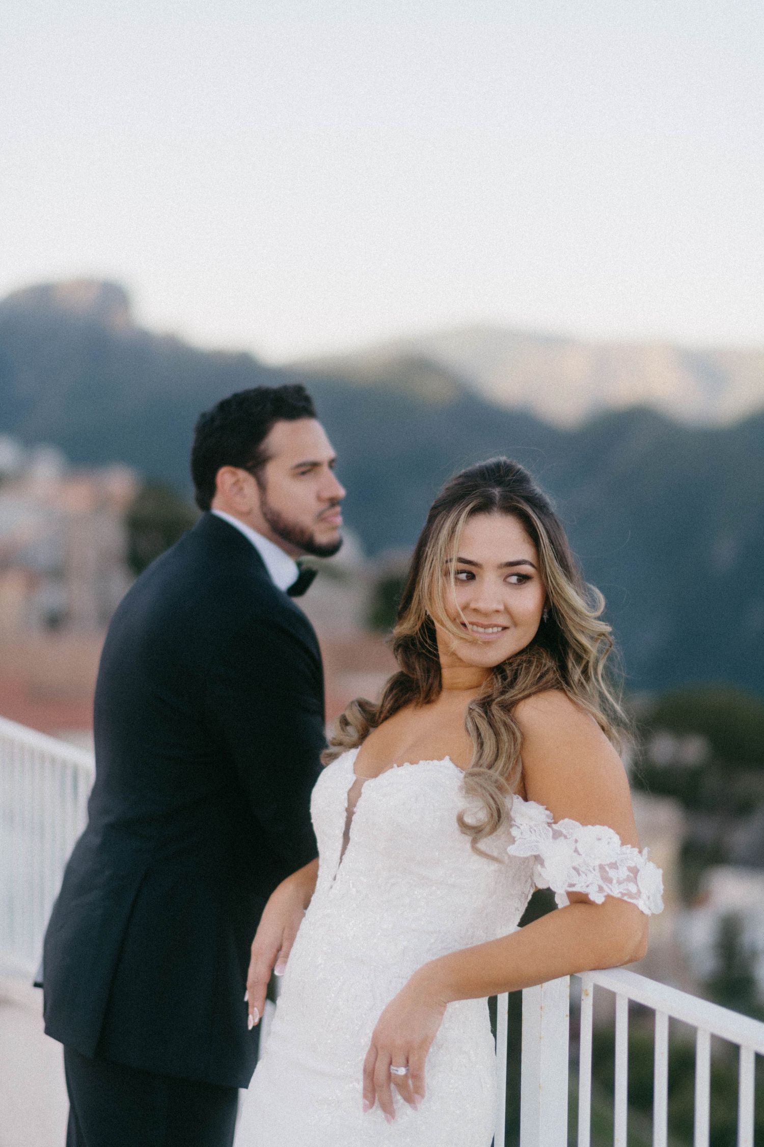 A couple in wedding attire pose on a balcony overlooking mountains and buildings during golden hour.