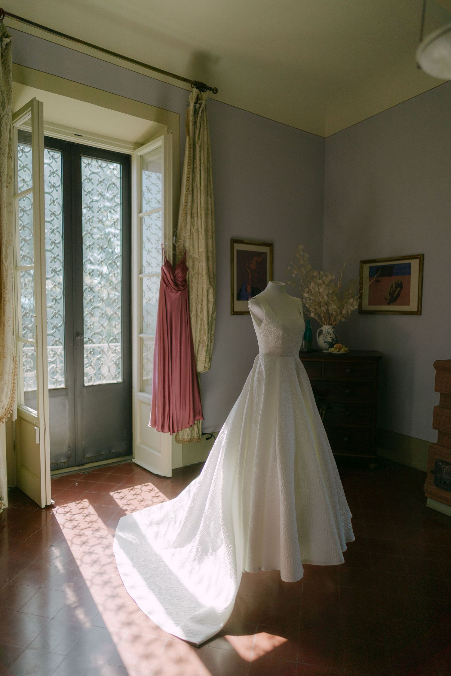Three white wedding dresses displayed in a sunlit room with hardwood floors and cream curtains.