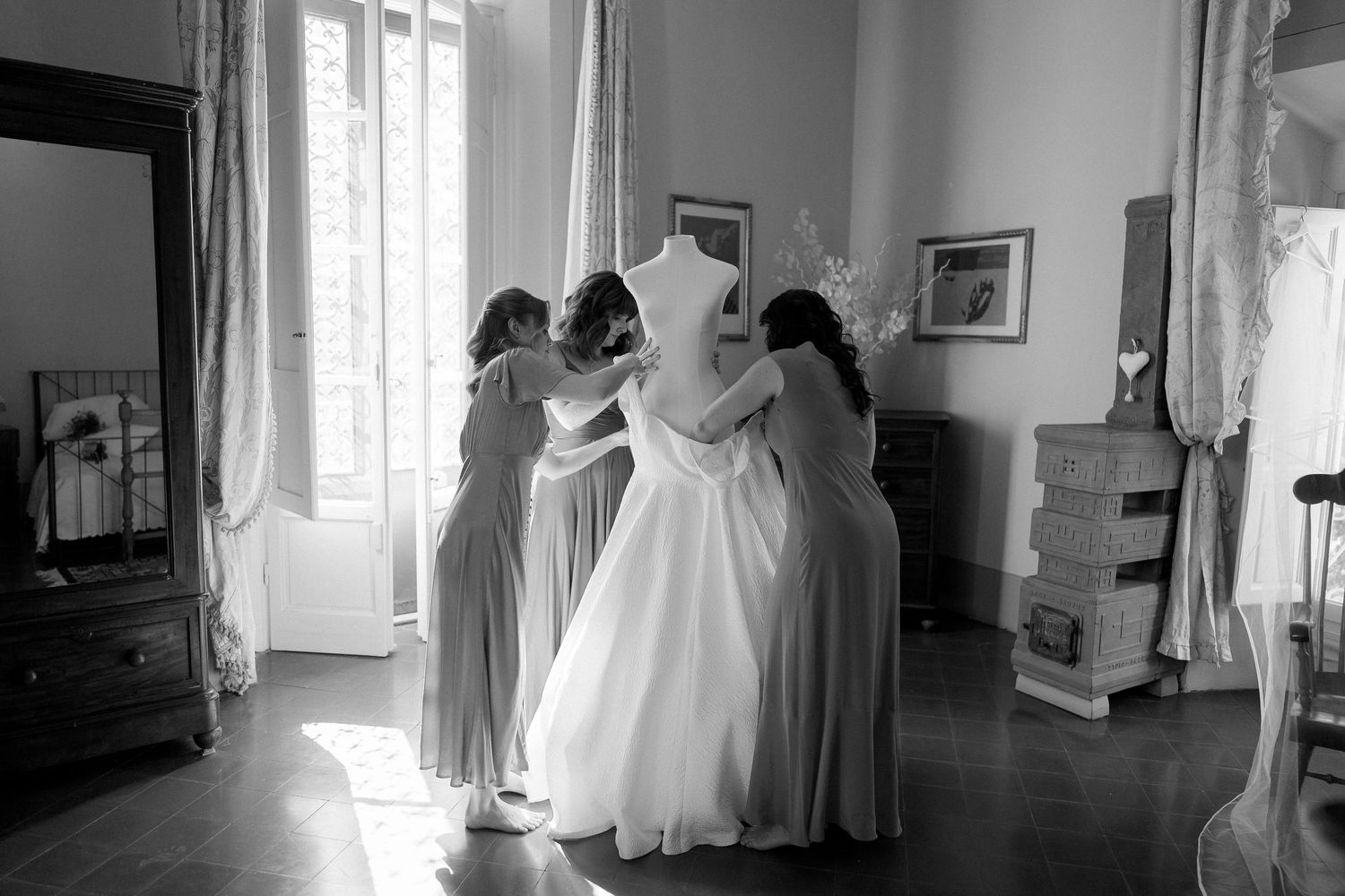 Black and white photograph showing a bride getting ready in a sunlit room with white curtains and elegant decor.