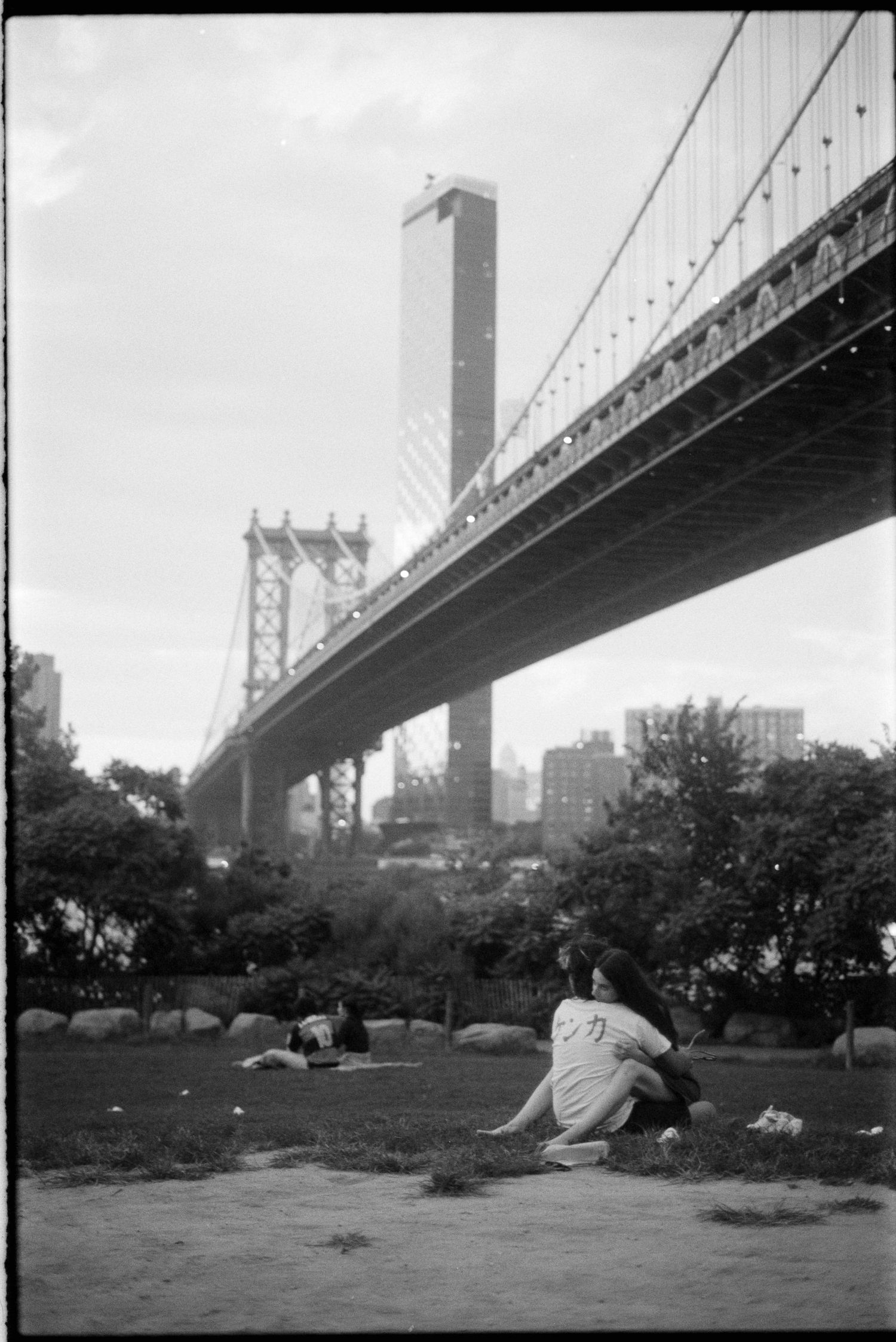 Black and white panoramic sequence showing people relaxing in a park beneath the Manhattan Bridge on a sunny day.