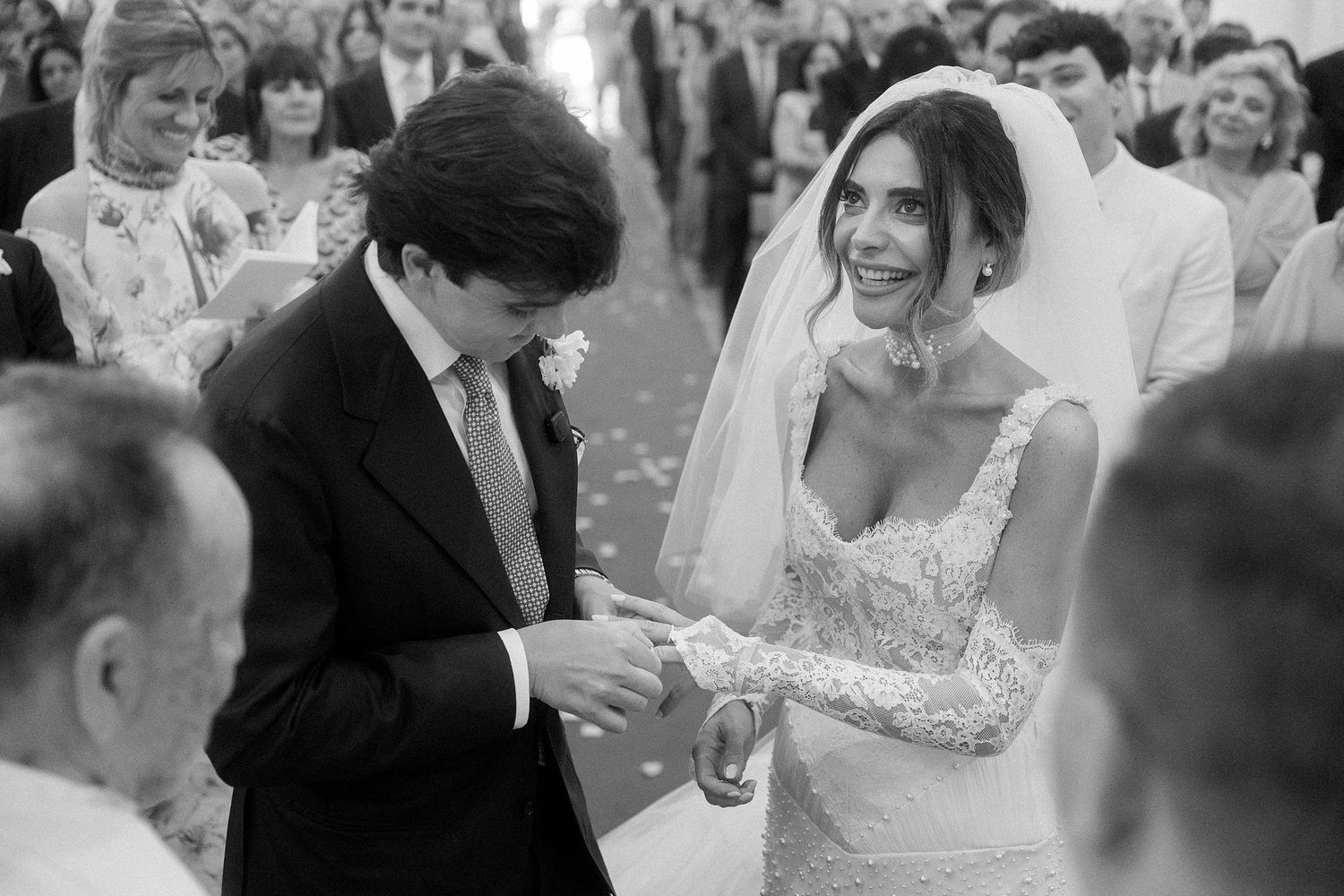 Black and white wedding ceremony photo of bride in lace gown and veil exchanging rings with groom outdoors.