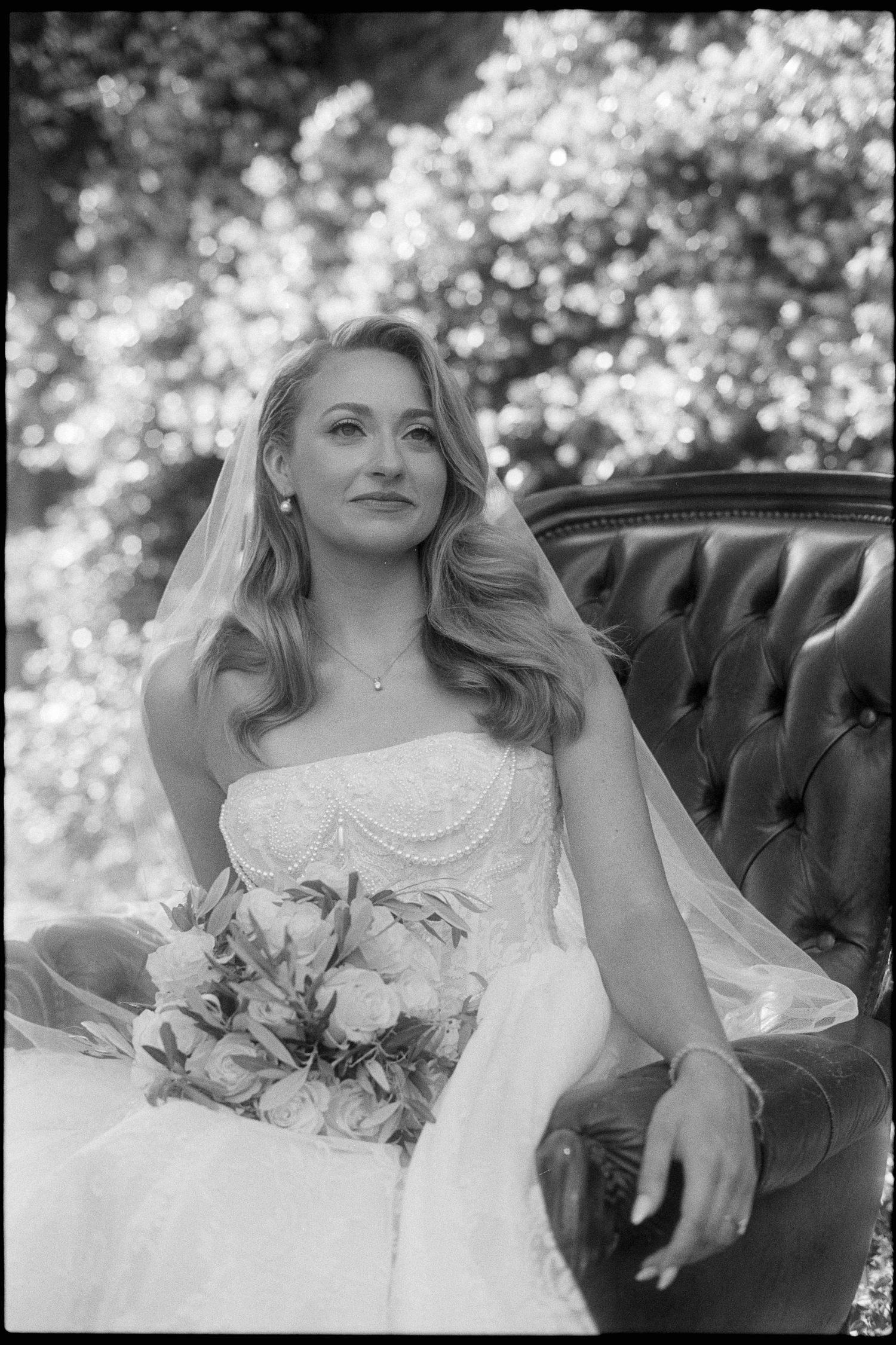 Black and white bridal portrait of a blonde bride in a white gown holding a bouquet, seated on a leather chair outdoors.