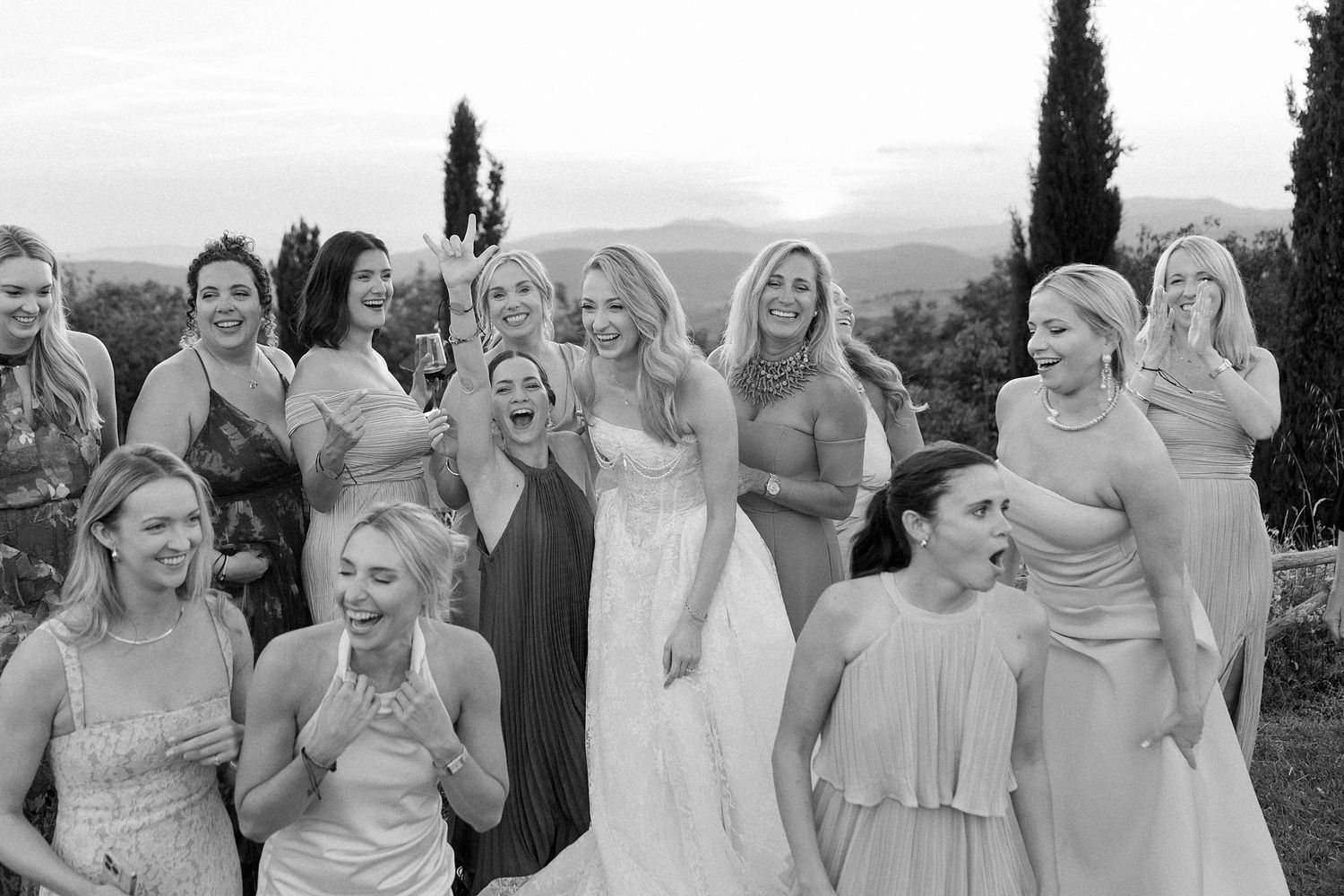 Black and white photo of a bride and bridesmaids laughing joyfully outdoors with scenic hills and cypress trees behind them.