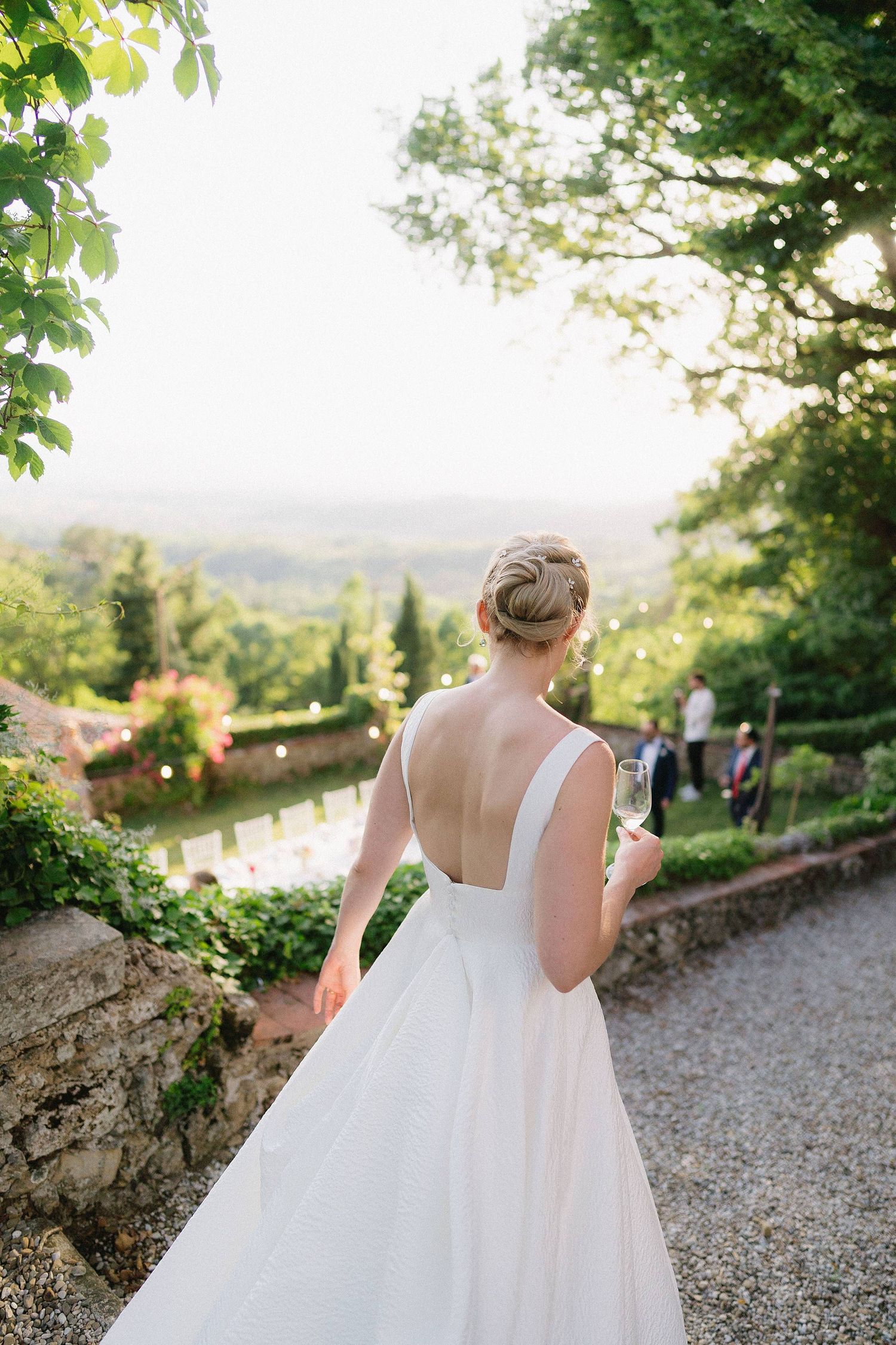 A bride in a white wedding dress holds a glass while overlooking a scenic Tuscan landscape.
