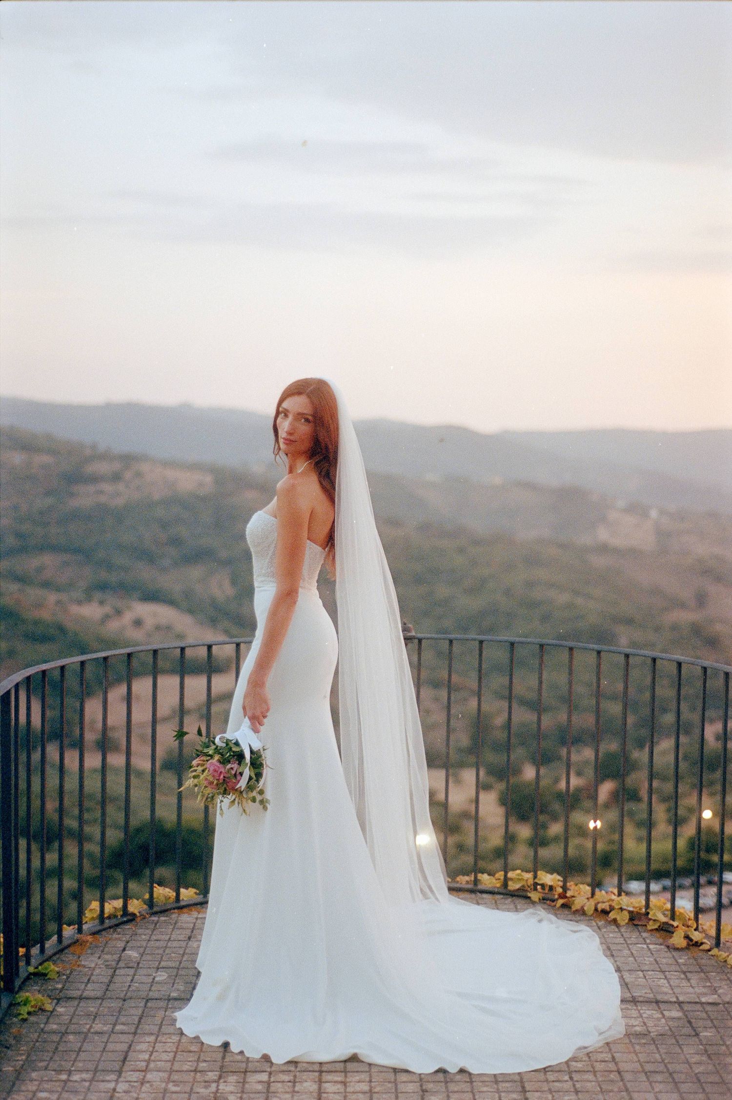 A bride in a flowing white gown stands on a scenic mountain overlook terrace during sunset.
