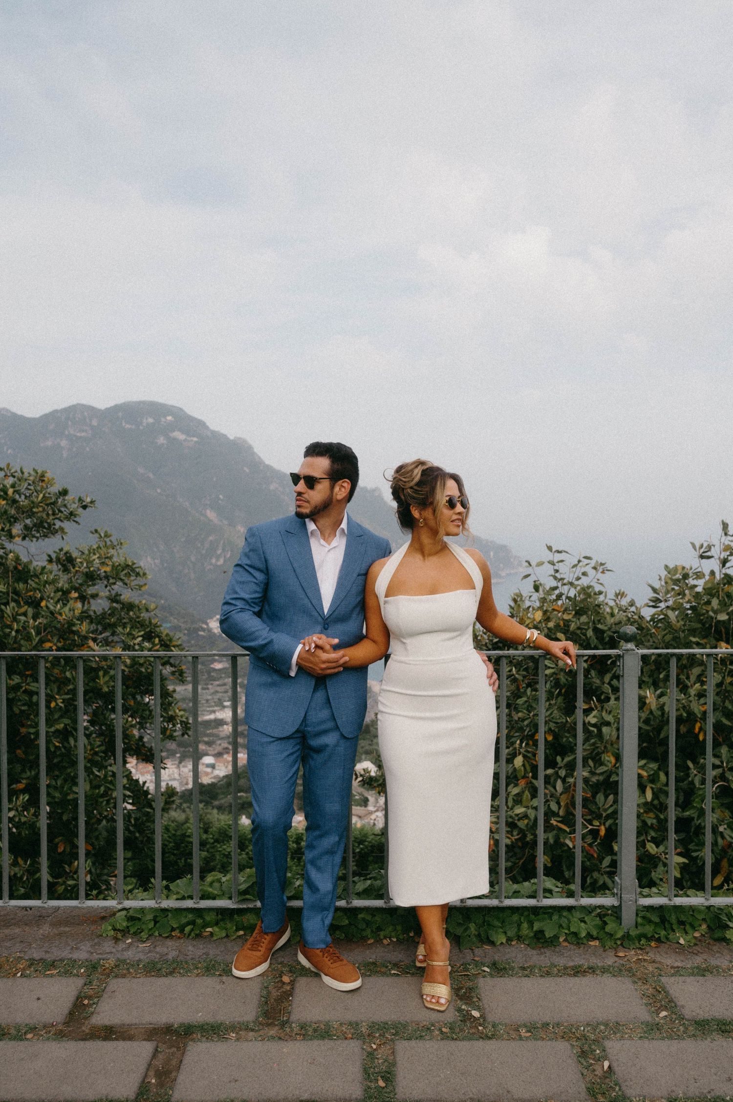 A couple in formal attire poses on a scenic mountain overlook with a coastal view in the background.