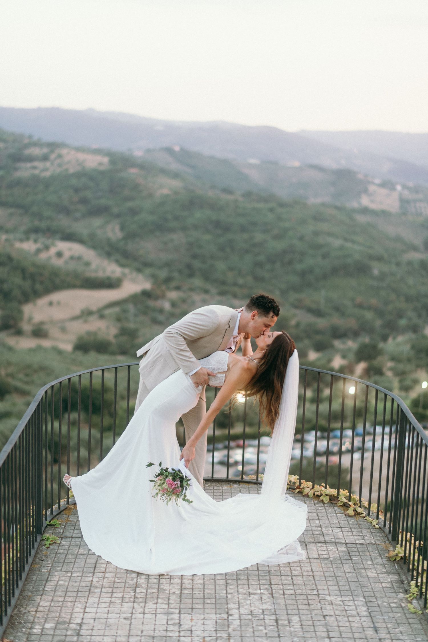 Romantic wedding couple dips and kisses on a scenic mountain overlook with dramatic valley views in the background.