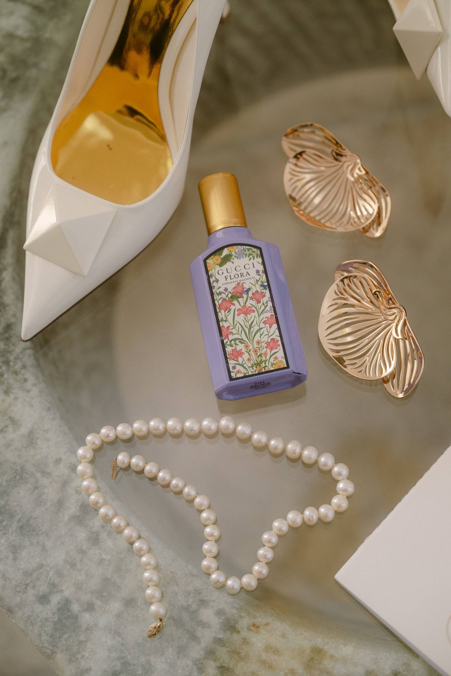 White high heel shoes with pearl necklace and gold earrings arranged on gray velvet surface with perfume bottles.