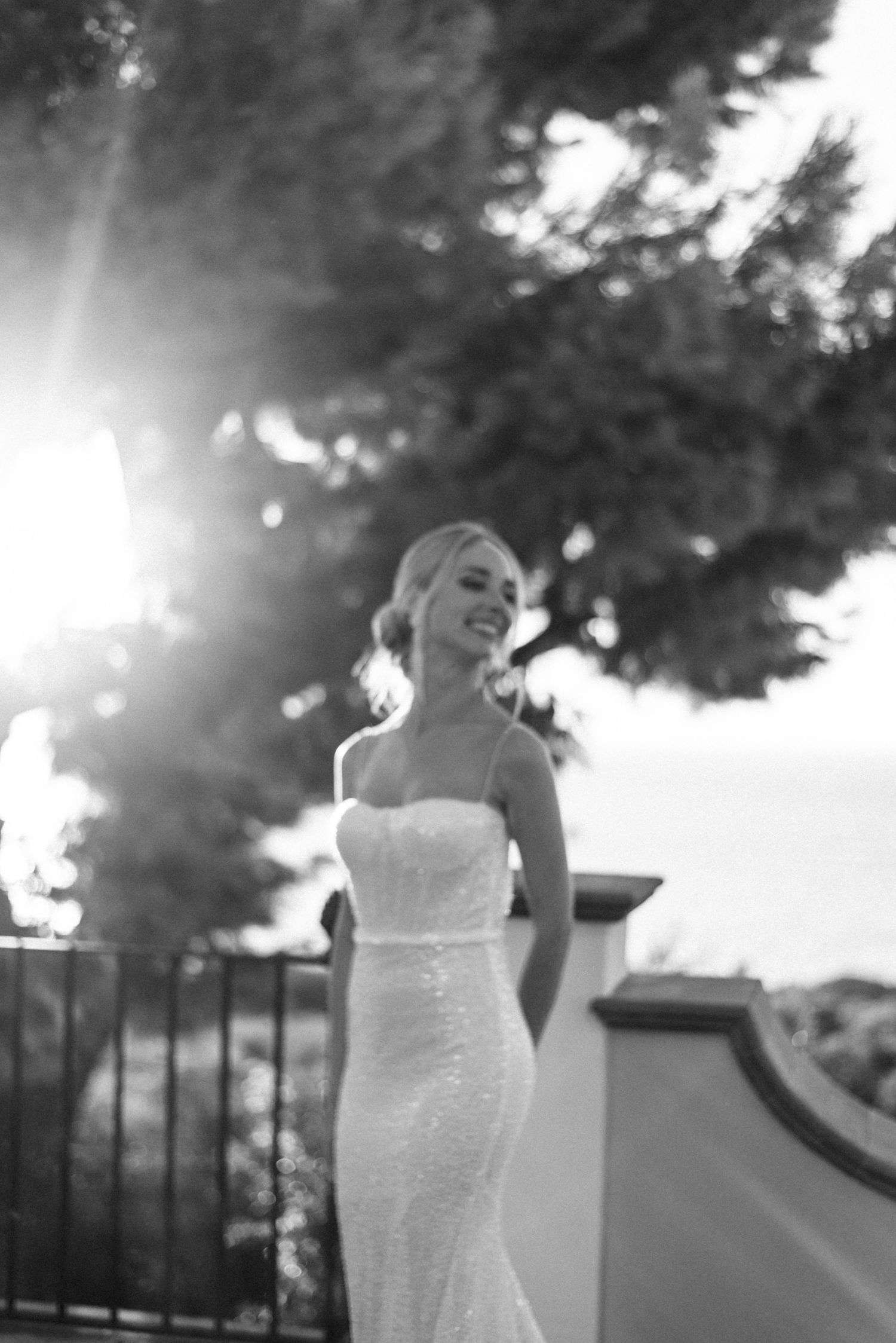 Black and white photo of a bride in a fitted white wedding dress standing in sunlight with trees in the background.