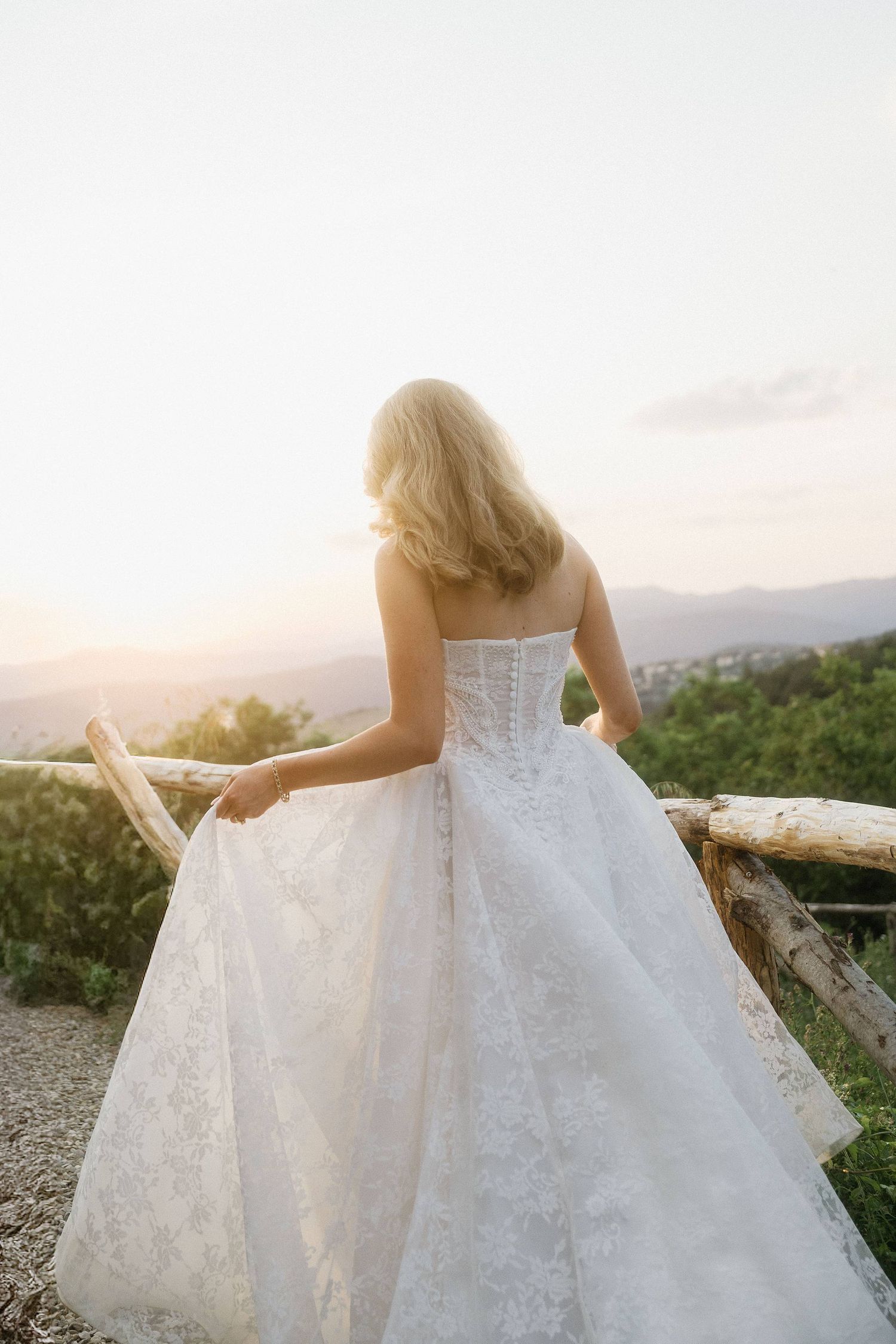 Bride in a strapless lace wedding gown stands at a wooden railing, gazing at a sunset landscape.