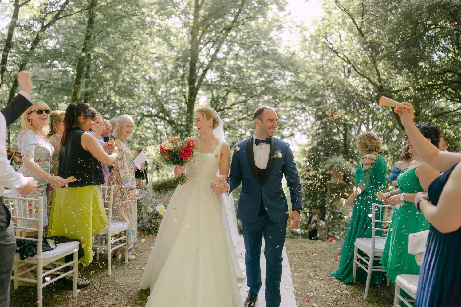 Newlyweds walk down outdoor aisle between white chairs as wedding guests celebrate in a wooded garden setting.