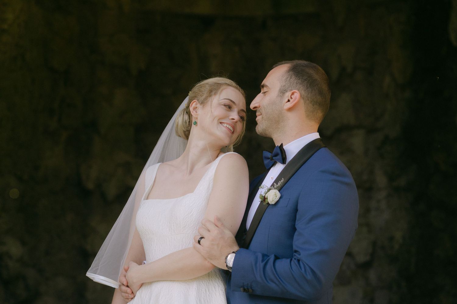 A bride and groom share an intimate moment against a dark stone wall during their wedding celebration.
