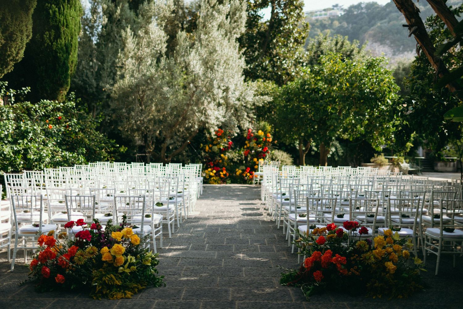 White garden chairs arranged for an outdoor wedding ceremony with colorful floral arrangements and mountain views in background.