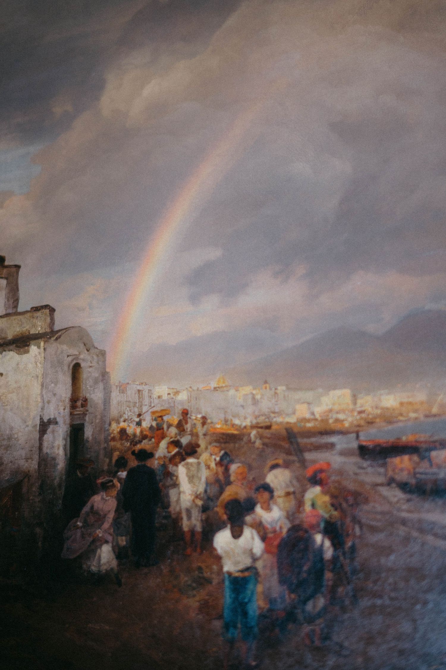 A crowd of people gather under a rainbow in a moody coastal scene with stormy clouds and white buildings.