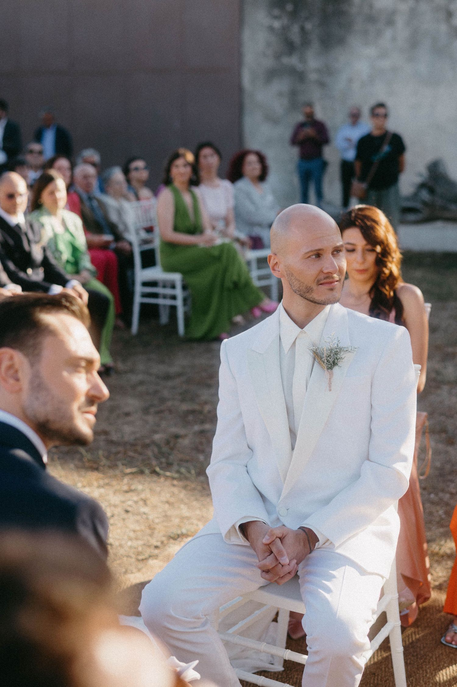 Wedding guests seated outdoors at a rustic ceremony venue with white chairs and natural lighting.
