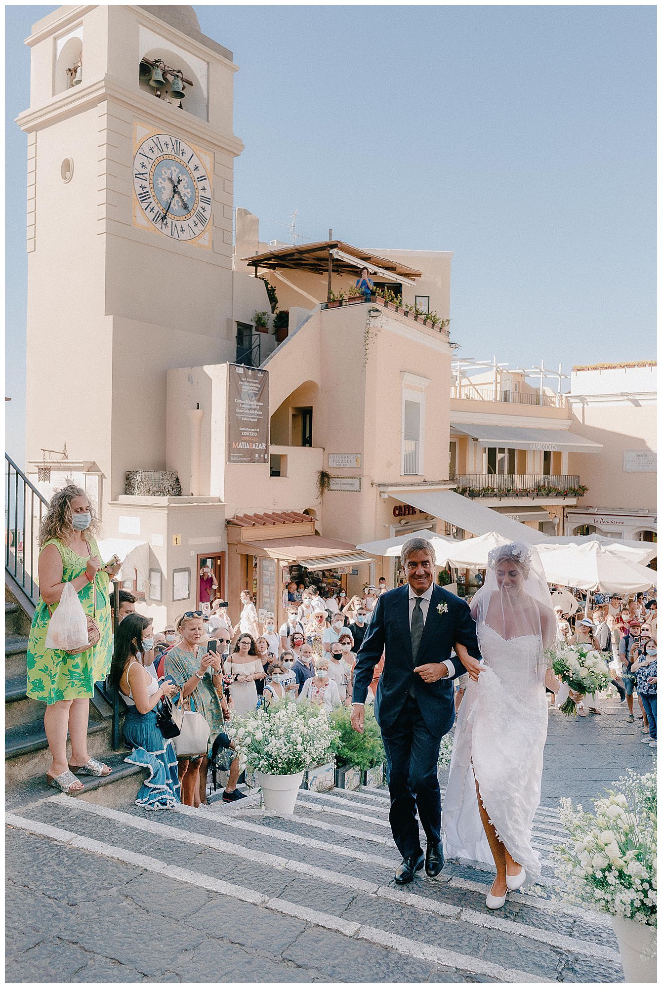 A scenic wedding ceremony takes place in front of a historic clock tower in a Mediterranean coastal town square.