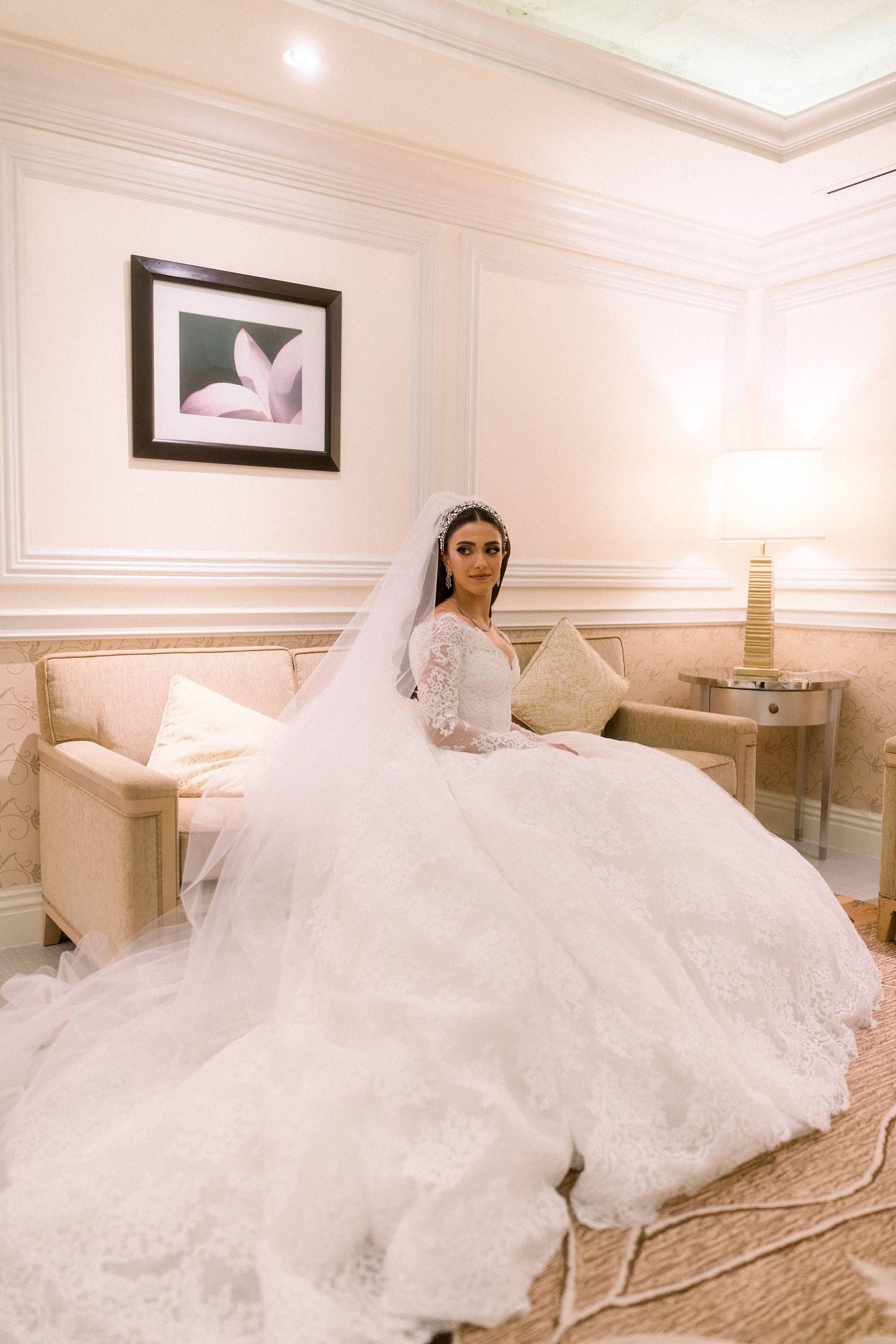 A flowing white wedding dress with intricate lace details spreads across a luxurious beige sofa in an elegant room.