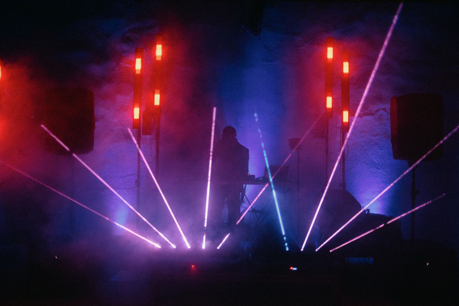 Colorful laser beams and red lights illuminate a dark concert stage with dramatic purple and blue atmospheric effects.
