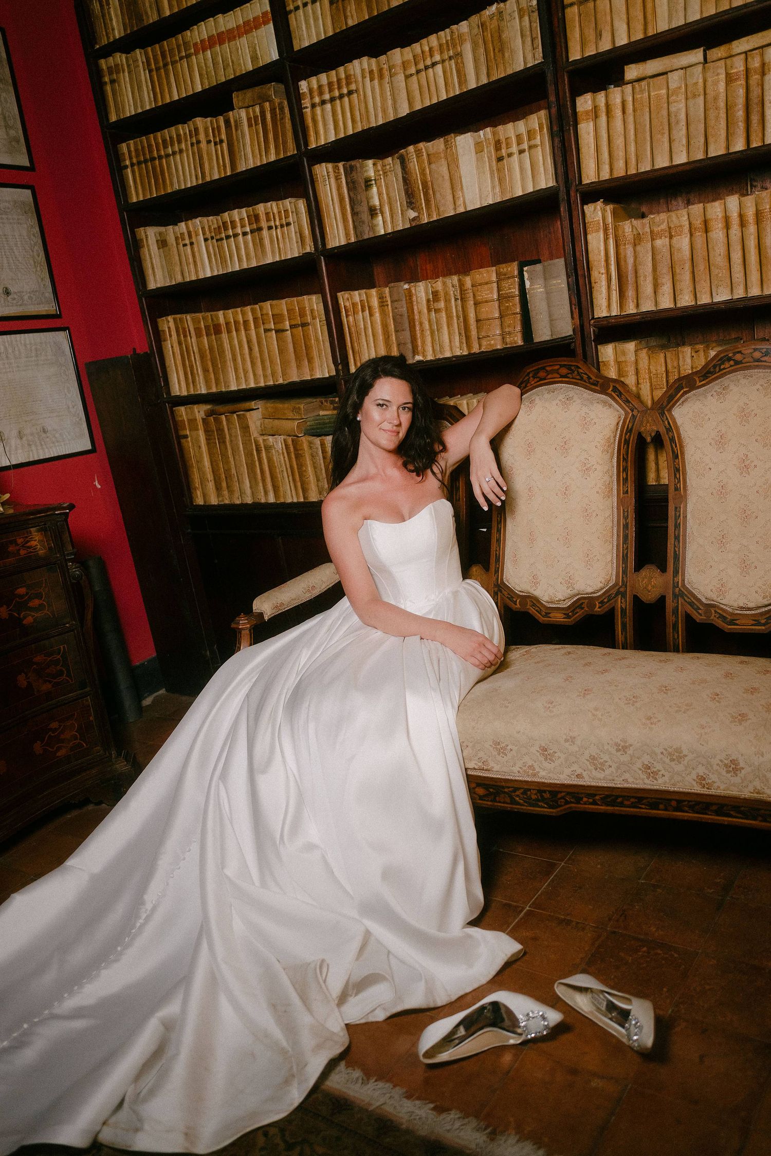 Elegant bride in white satin gown poses on vintage chaise lounge in library with floor-to-ceiling bookshelves.