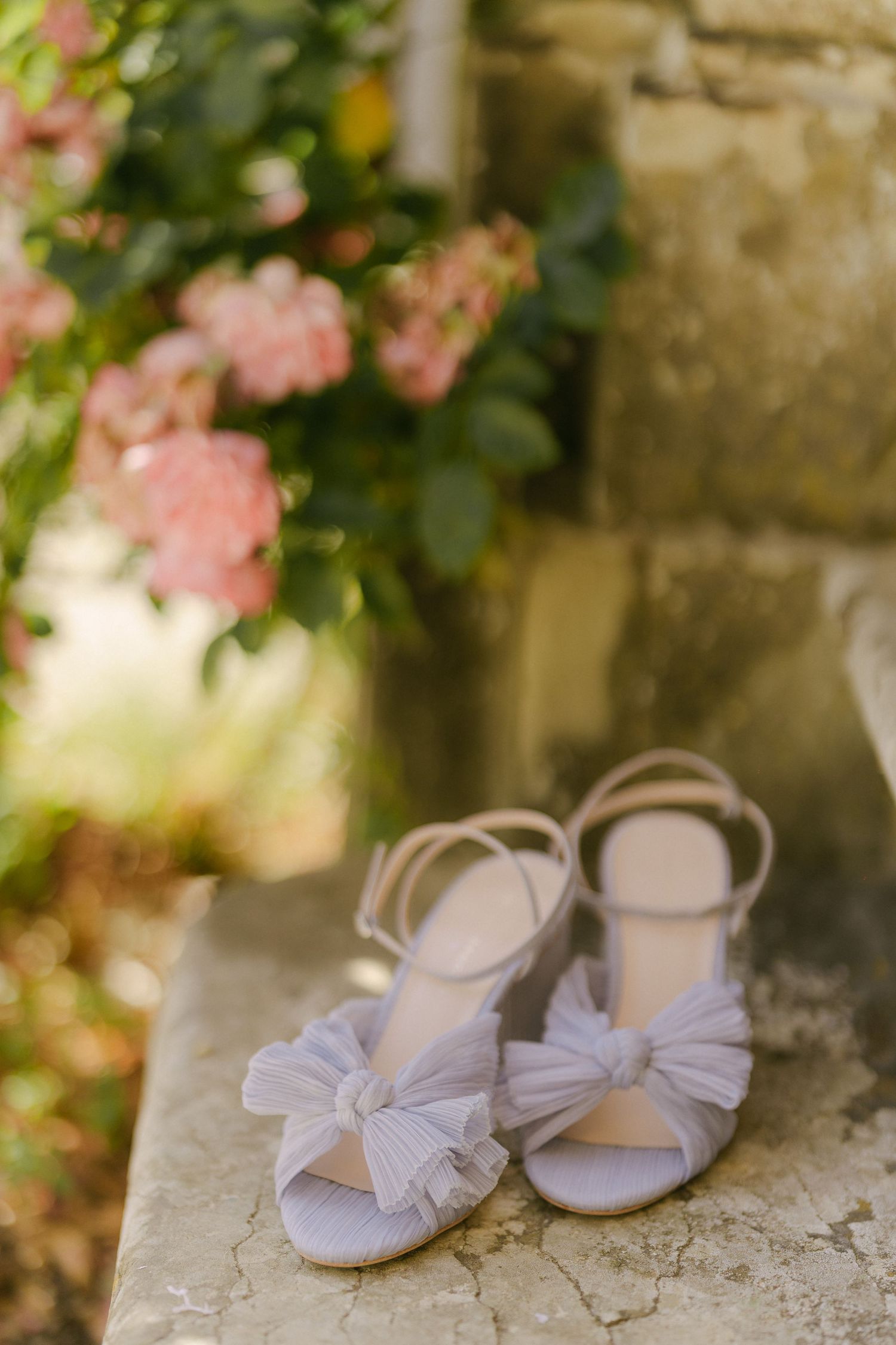 Elegant lavender bridal sandals with bow details sit on stone ledge with pink roses blooming in background.