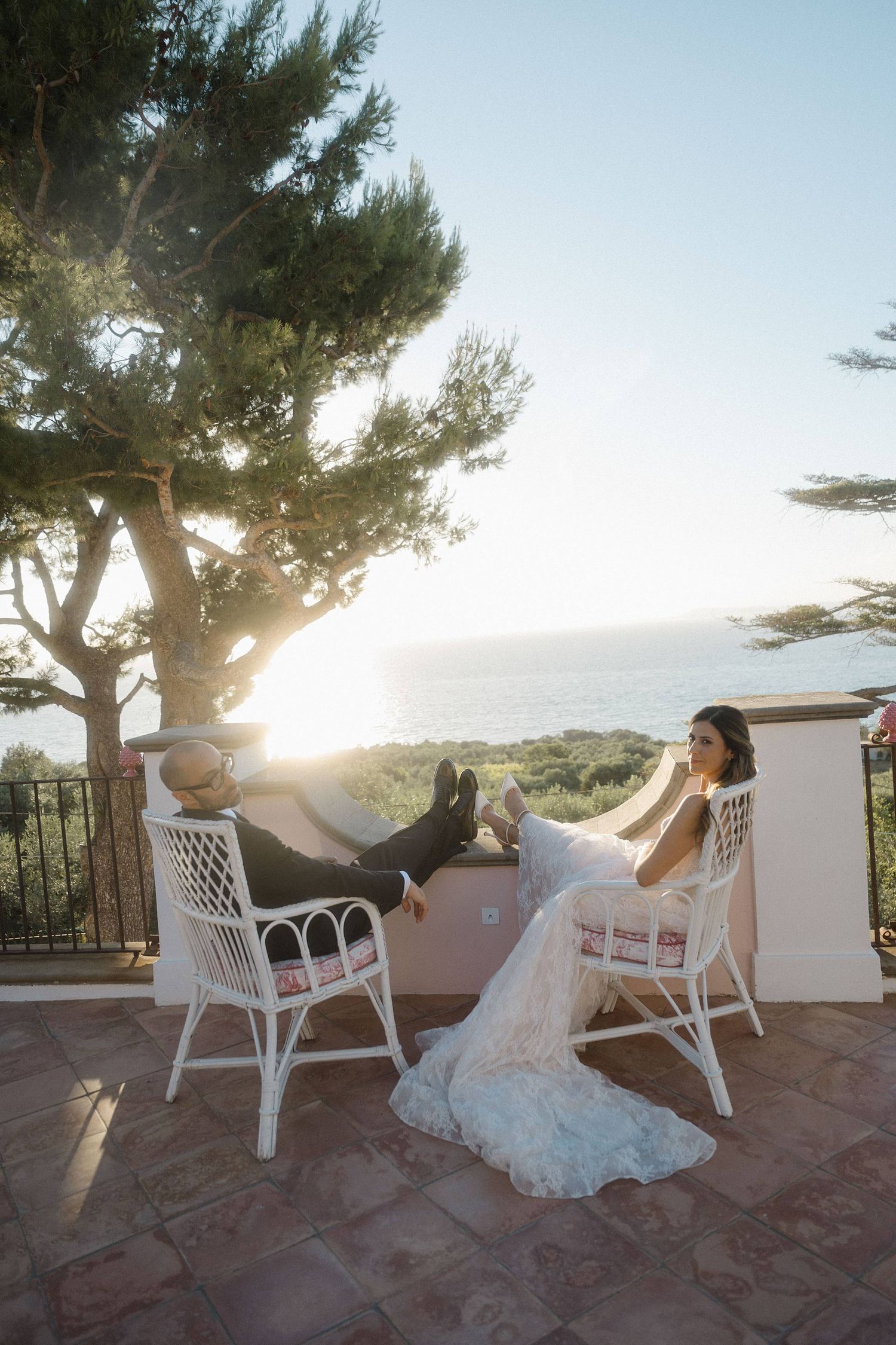Bride and groom relaxing in white wicker chairs on a terrace at sunset with ocean views behind them.