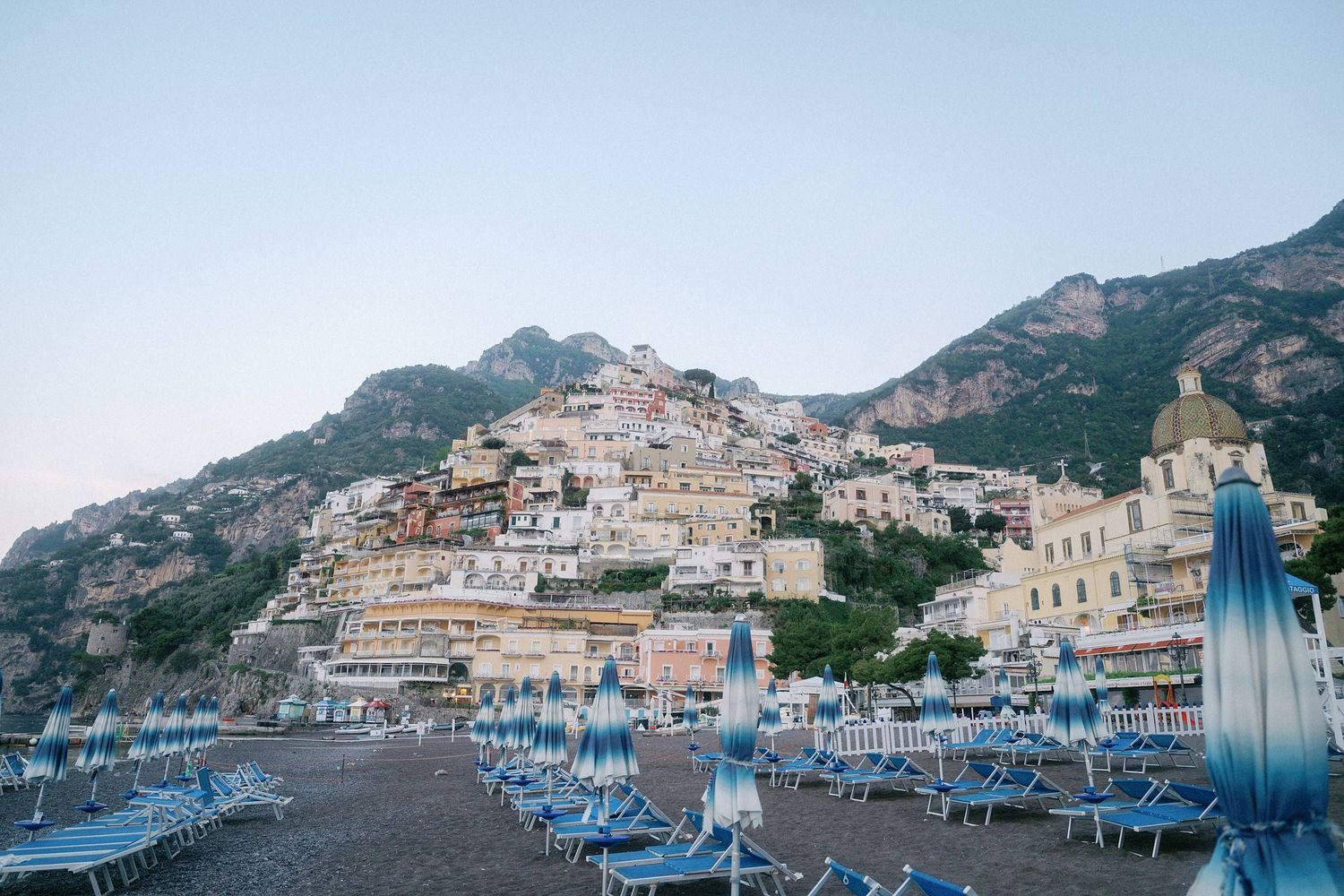 Colorful beach umbrellas and lounge chairs line the shore beneath the cascading hillside buildings of Positano, Italy.