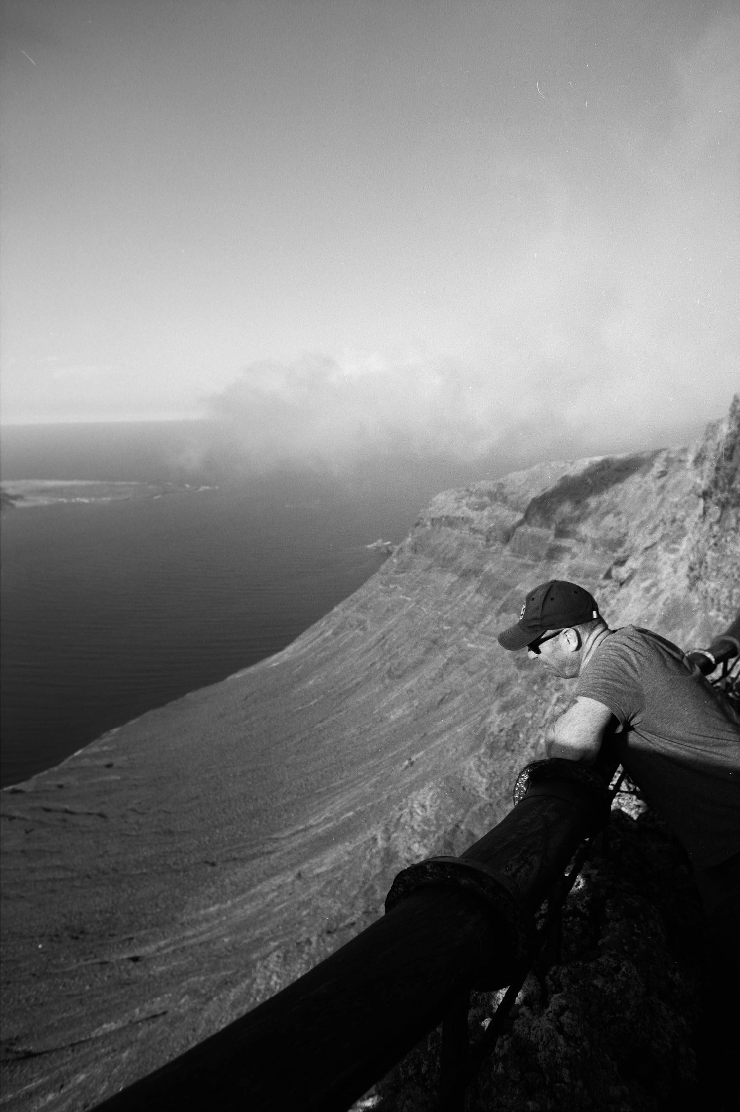 A hiker peers over the edge of a dramatic coastal cliff overlooking misty ocean views in black and white.