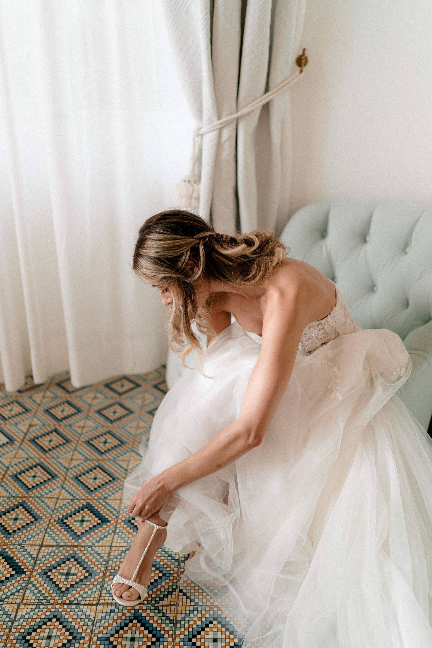 Bride in white wedding dress adjusts her strappy sandal while seated on a blue tufted couch.