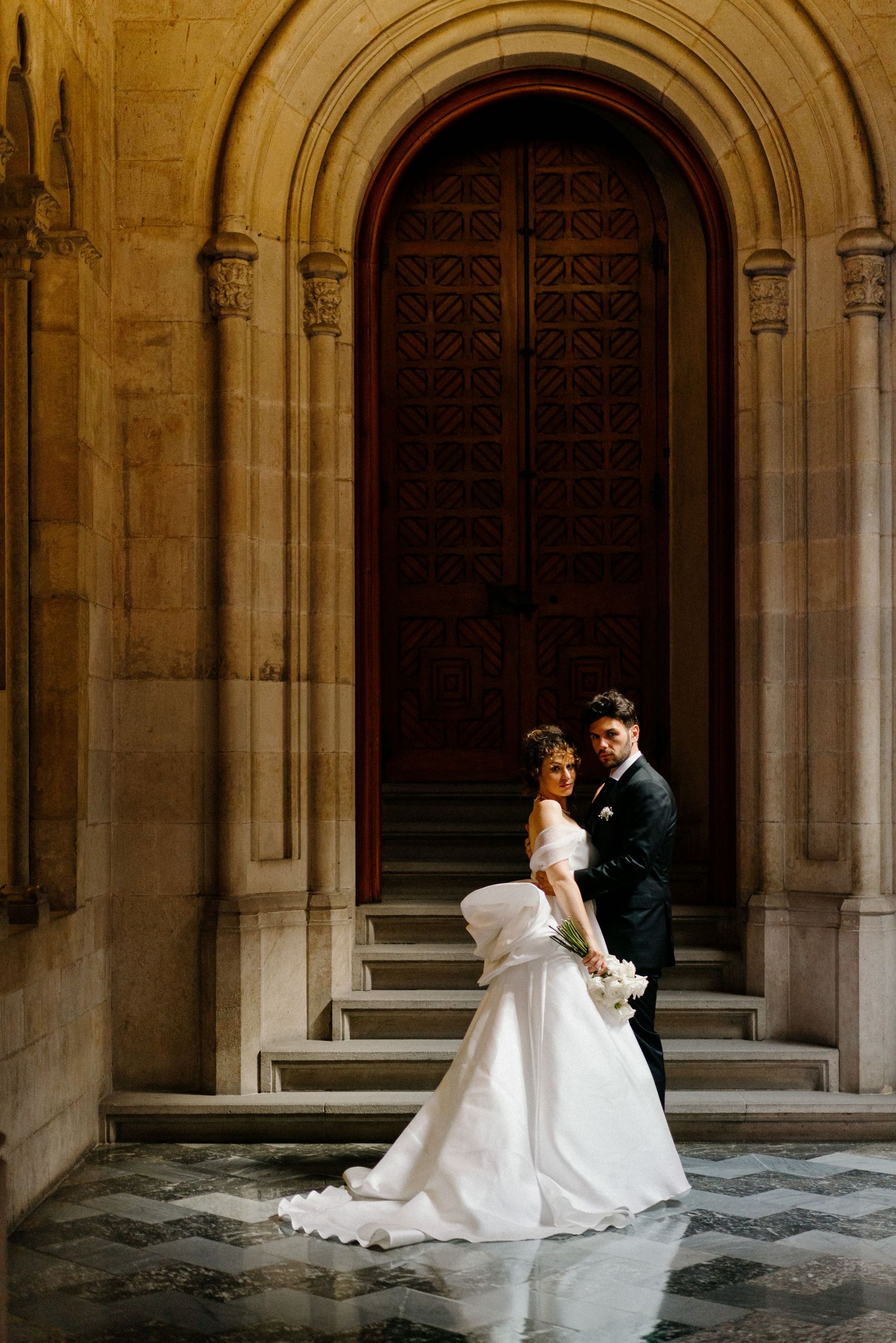 A couple stands elegantly on stone steps under a grand arched doorway at a historic cathedral-like building.