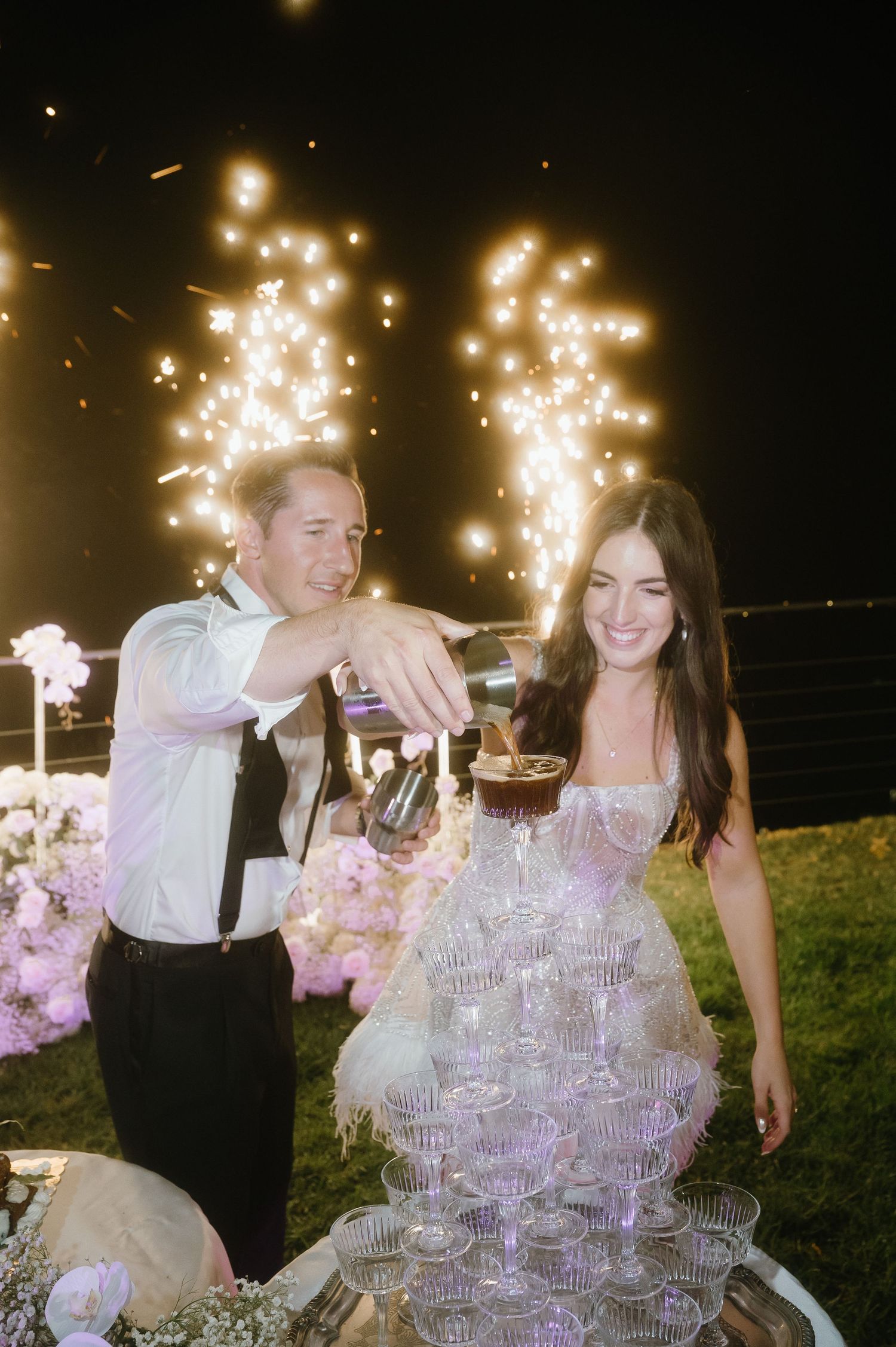 Couple pouring champagne into a tower of crystal glasses at an elegant outdoor evening celebration.