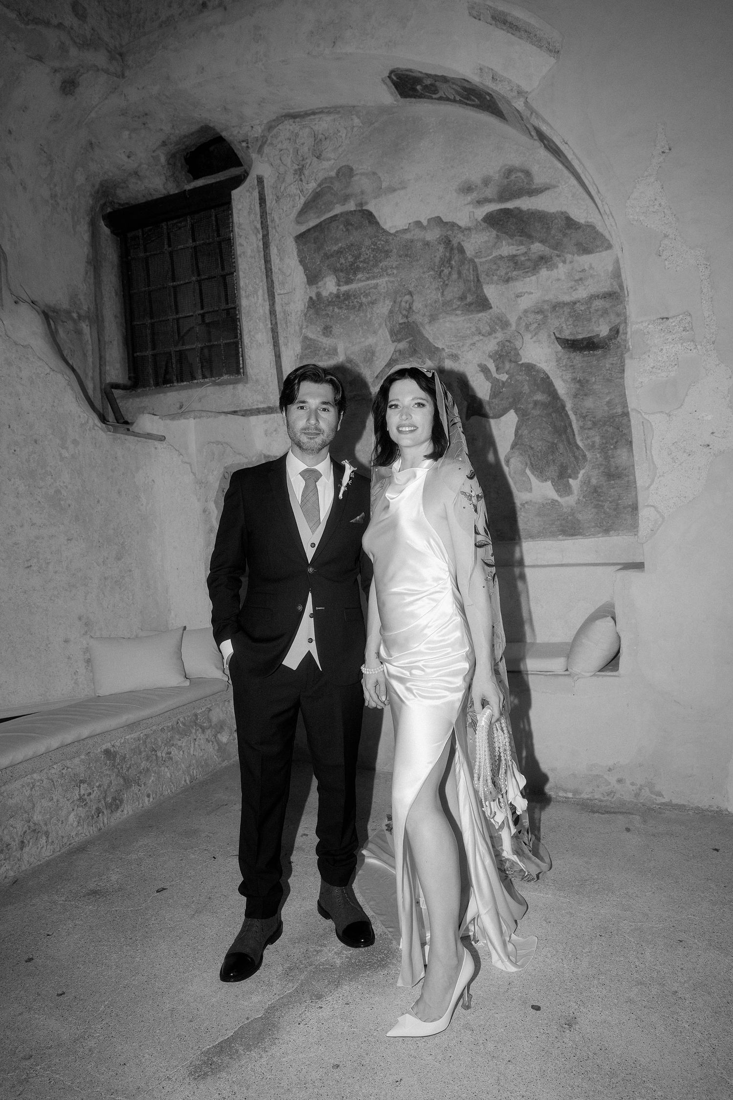 Black and white wedding photo of a couple posing against a rustic wall with vintage architectural details.