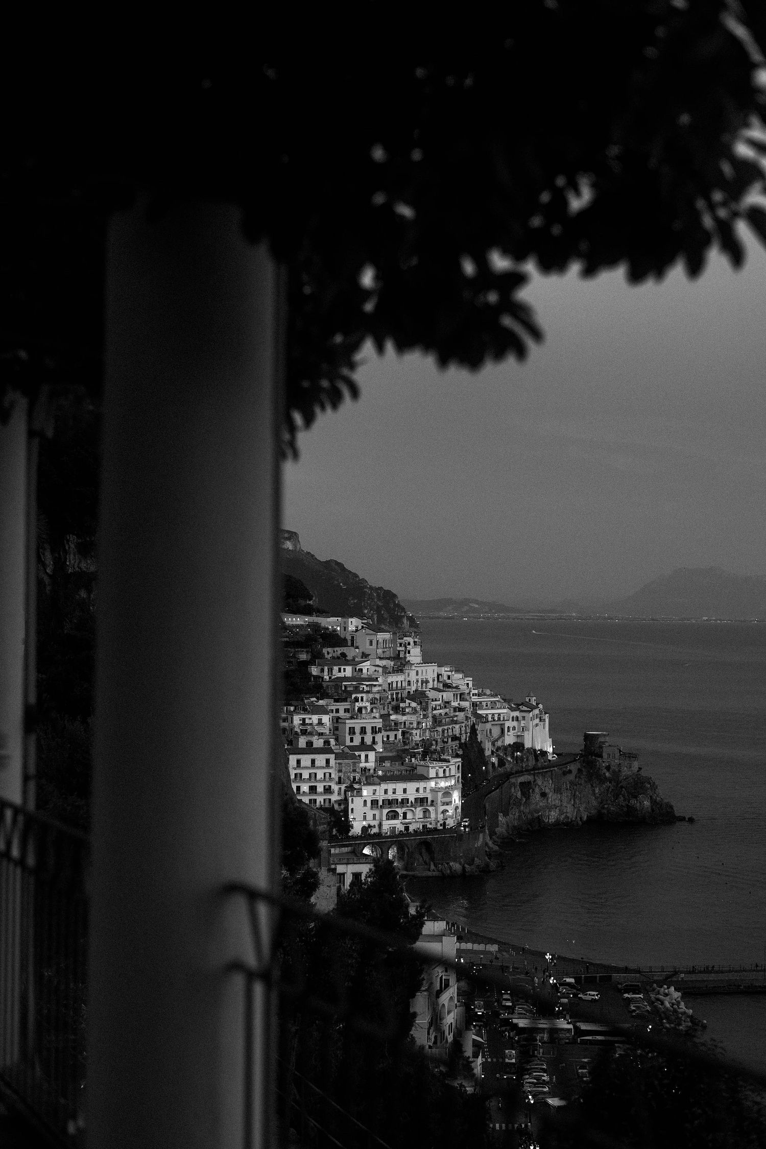 Black and white coastal landscape of Amalfi, Italy showing white buildings nestled on cliffside overlooking Mediterranean Sea.