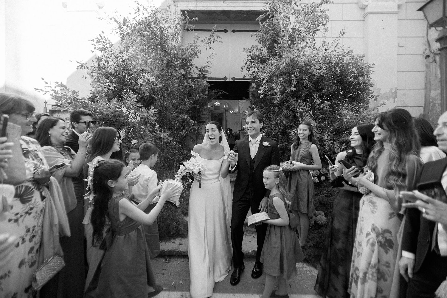 Black and white wedding photo of couple exiting ceremony, surrounded by cheering guests holding flowers outdoors.
