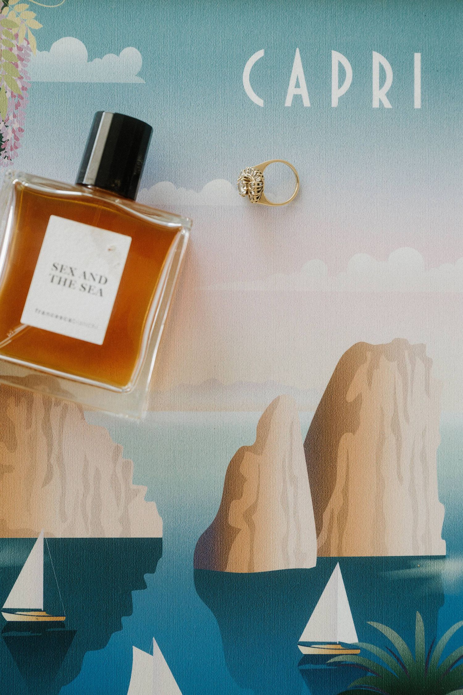 Perfume bottle of Sex and the Sea by Strangelove NYC beside a gold ring on a Capri travel poster.