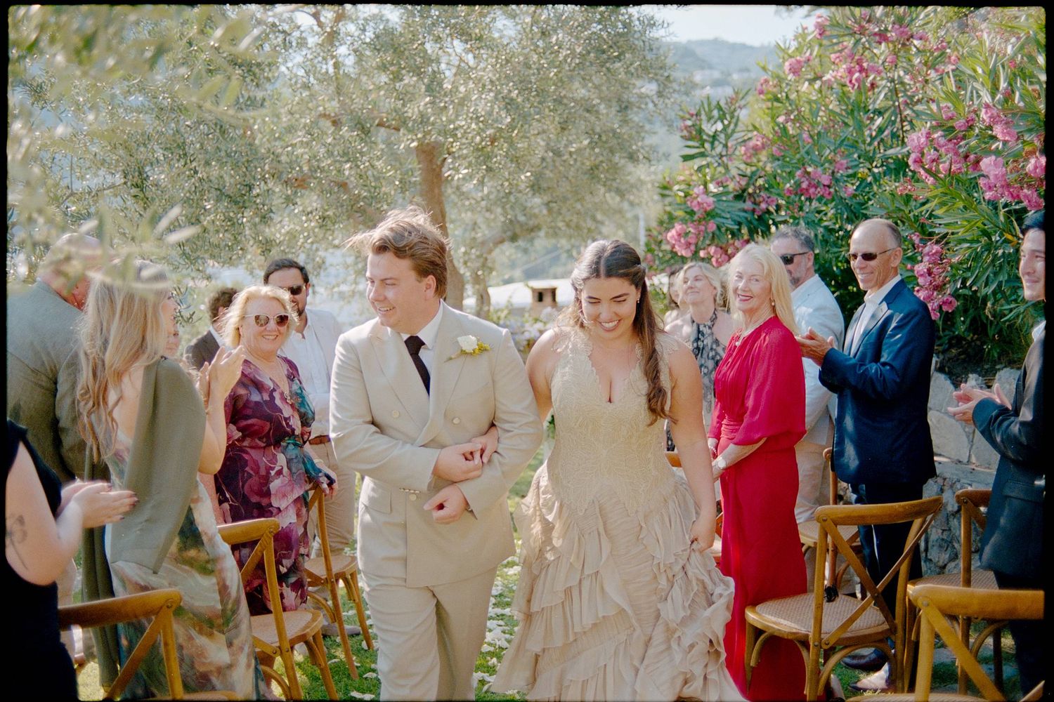 Bride and groom walk down the aisle at an outdoor wedding ceremony surrounded by guests and blooming flowers.