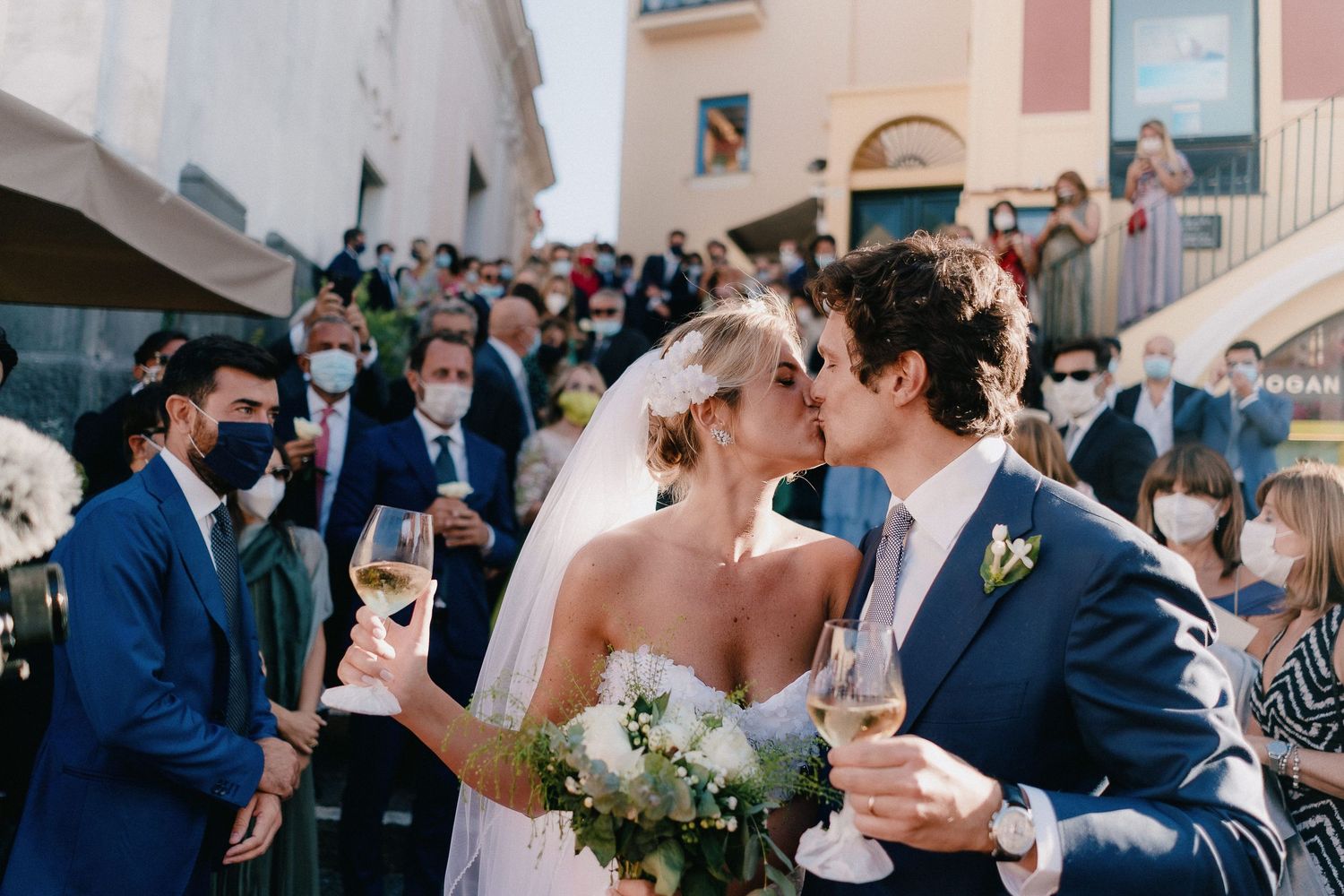 Newlywed couple sharing champagne toast with wedding guests at outdoor celebration during daytime.