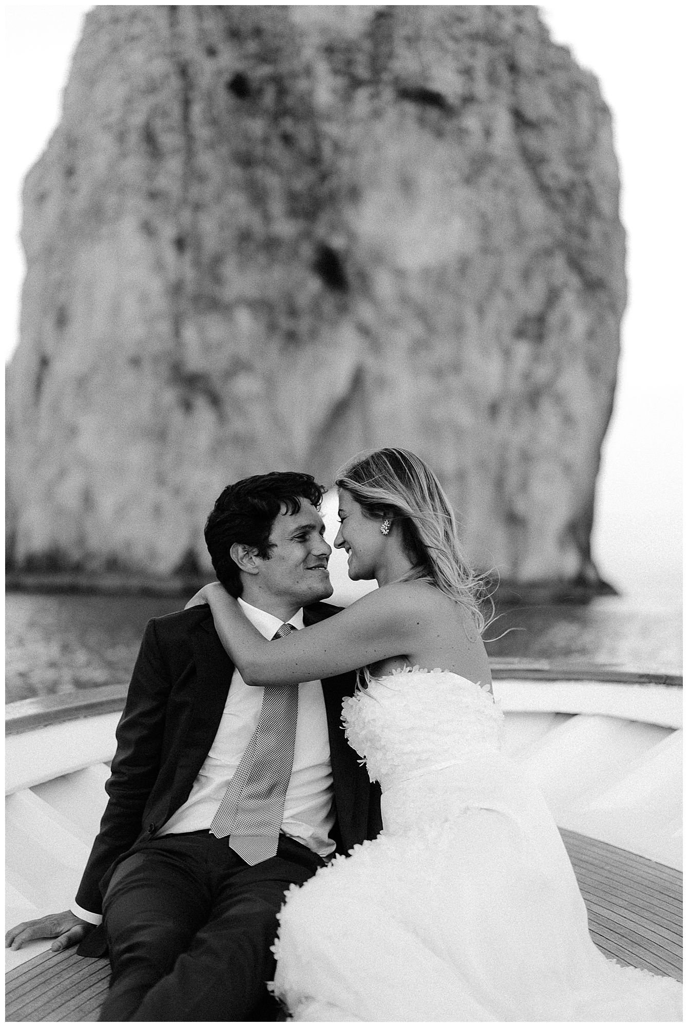 Romantic couple on boat embraces near towering coastal cliff in black and white photo.
